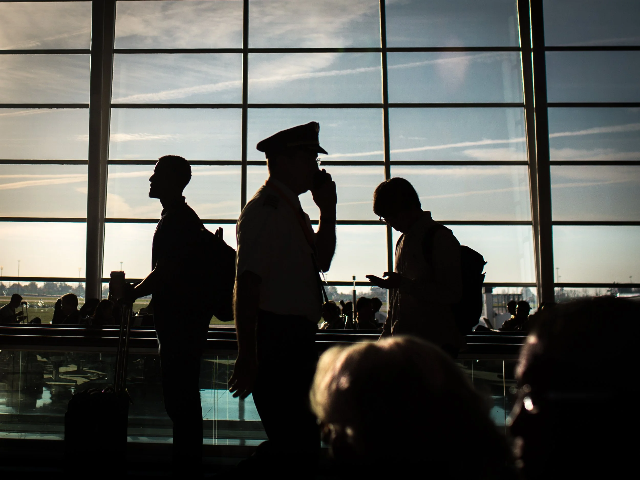 Silhouettes of three people, including a uniformed officer, inside an airport terminal with large windows and a view of the tarmac outside.