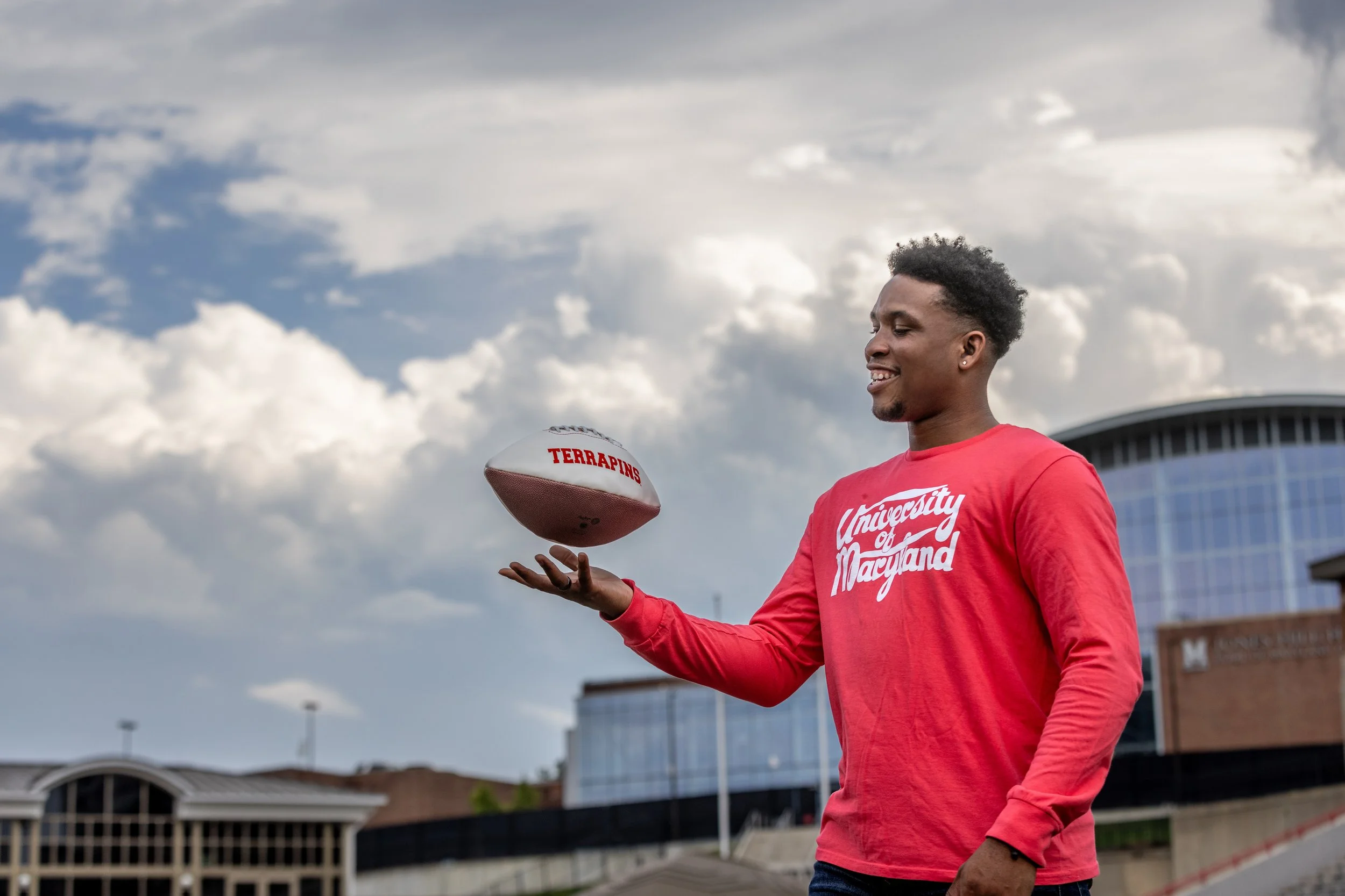 A young man in a red University of Maryland shirt is spinning a football with the word "Terrapins" written on it on his finger outdoors, with clouds and university buildings in the background.