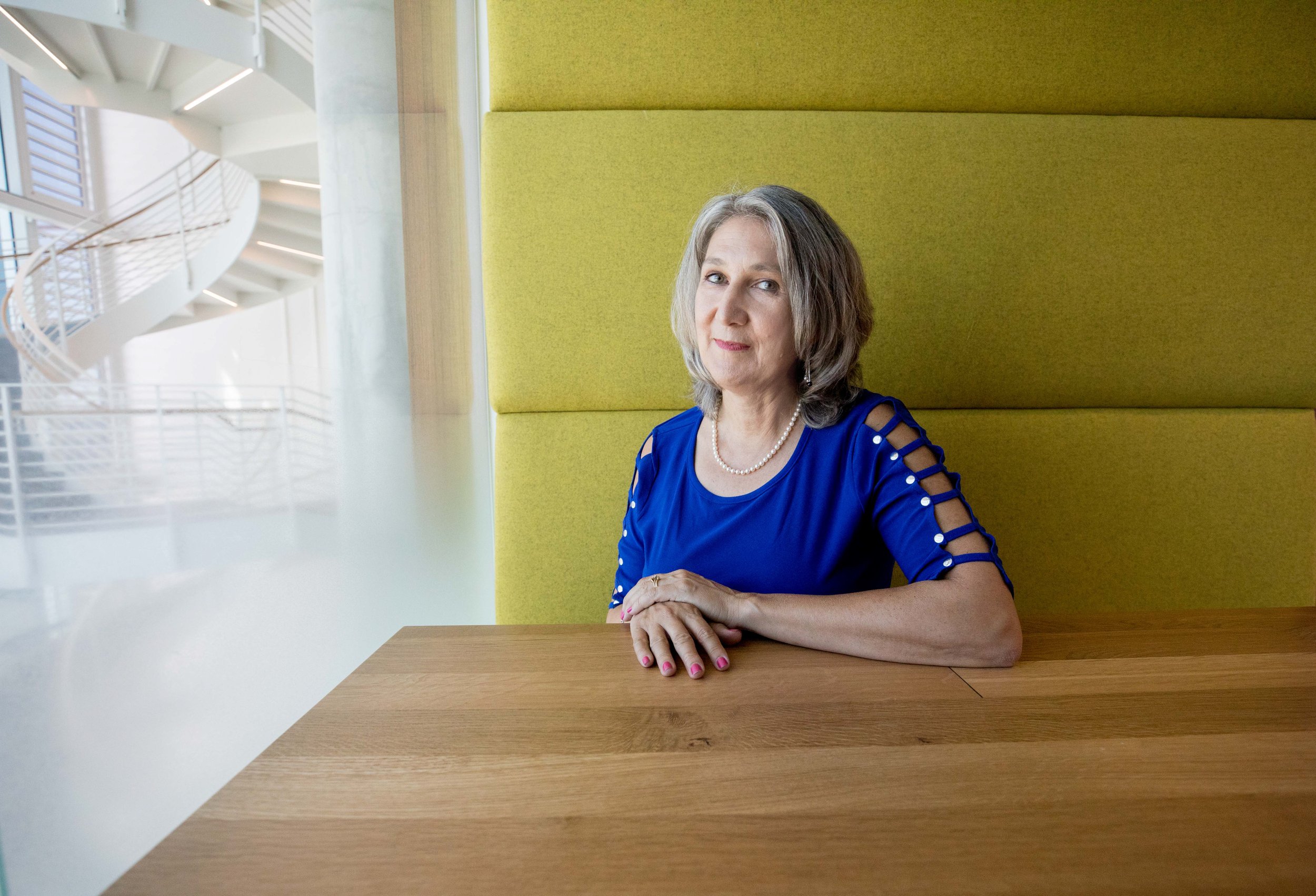 An older woman with gray hair sitting at a wooden table in a modern, brightly lit interior. She is wearing a blue dress with cut-out details on the sleeves and a pearl necklace. Behind her is a yellow cushioned wall and a large window showing a staircase and railing outside.