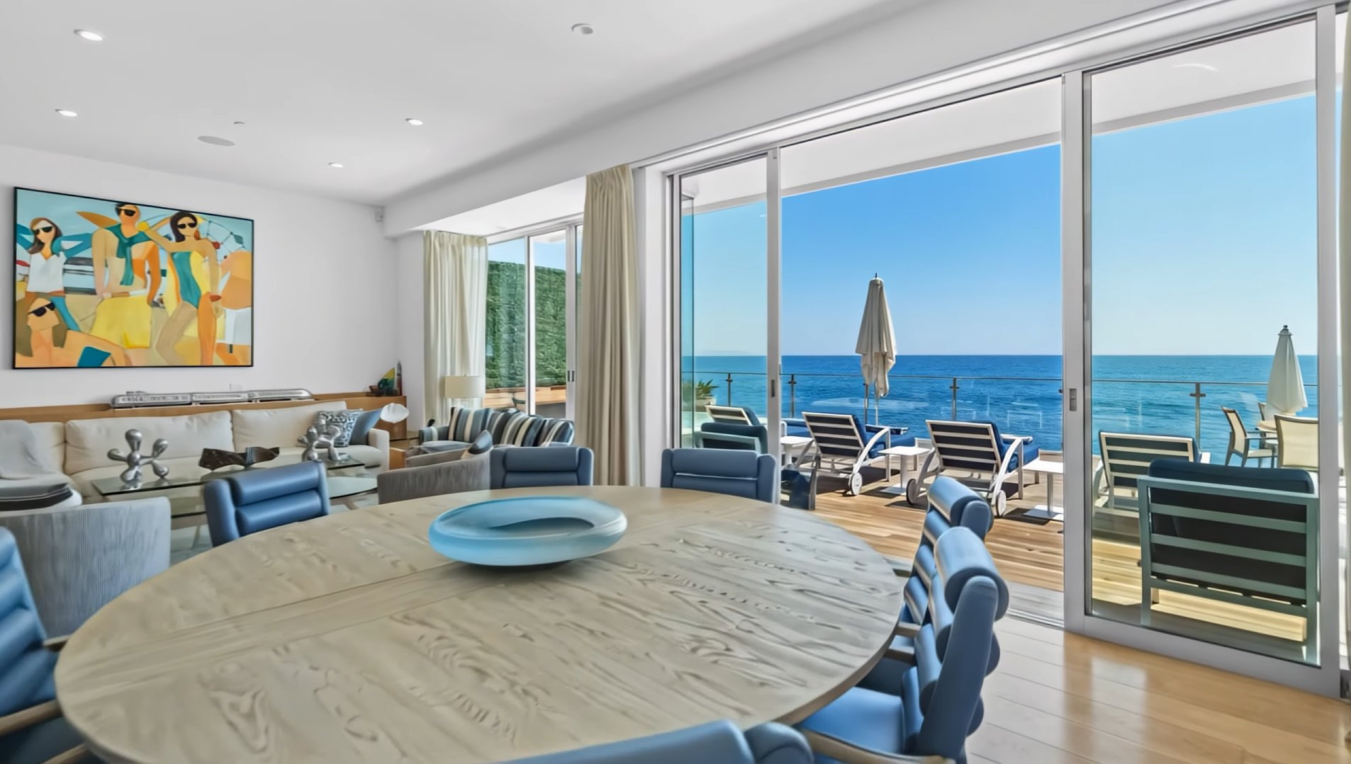 Living room with large glass sliding doors leading to a balcony overlooking the ocean, decorated with modern furniture, a round wooden table with blue chairs, a white sofa, and a colorful abstract wall art.