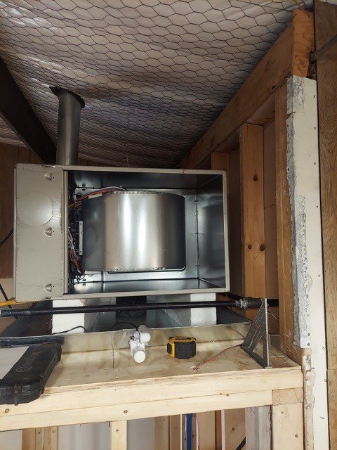 View of an HVAC furnace system installed in a wooden workshop, with a metal duct connection and some tools on the work surface.