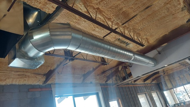 Silver industrial air duct running along the ceiling of a building with exposed insulation and window.
