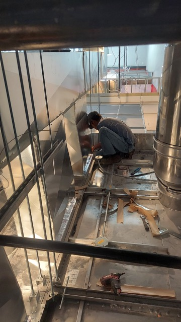 A worker crouched down assembling or repairing a metal structure in a construction or maintenance area with tools and materials scattered around.