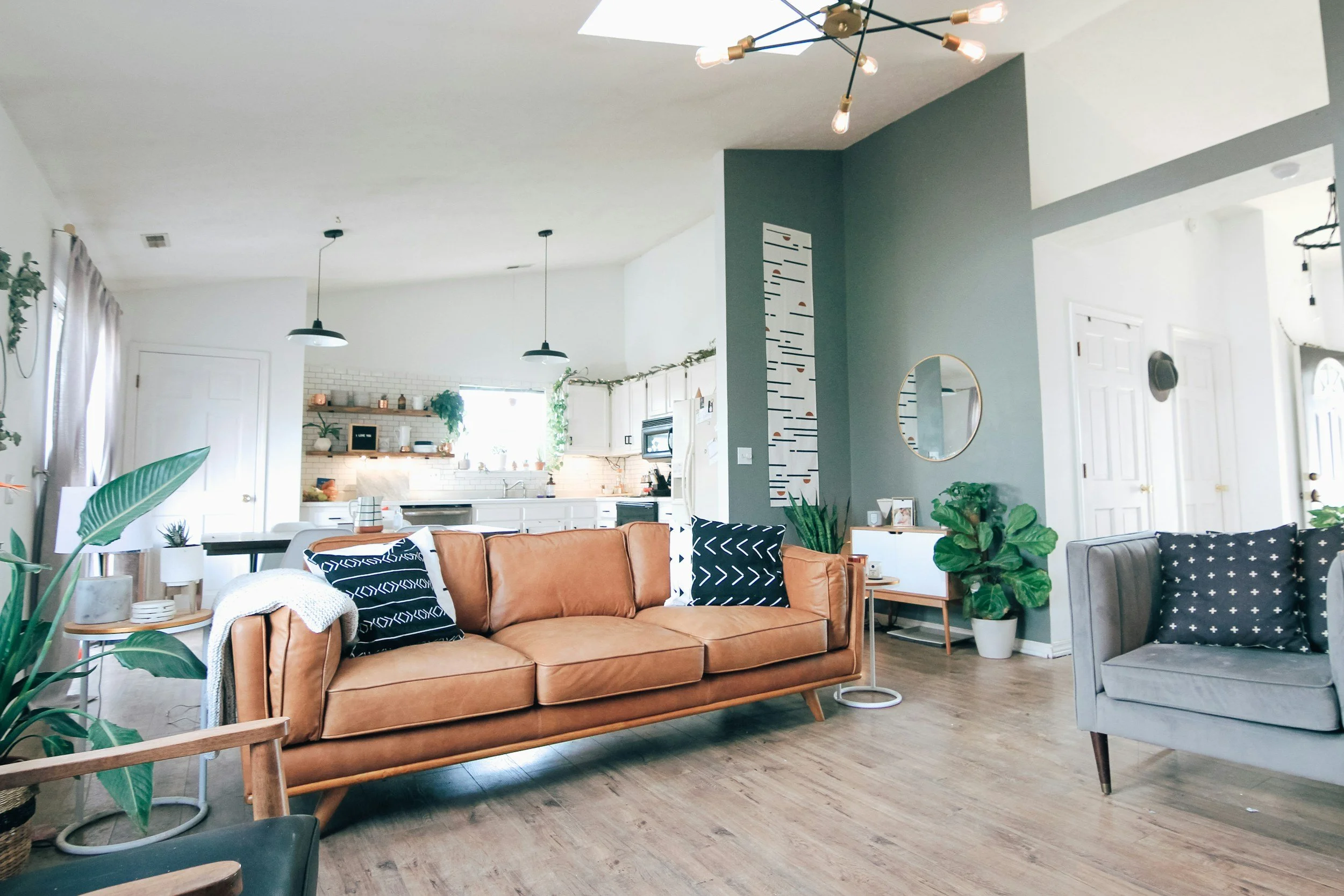 Living room with brown leather sofa, gray armchair, plants, and modern decor, open to kitchen in background.
