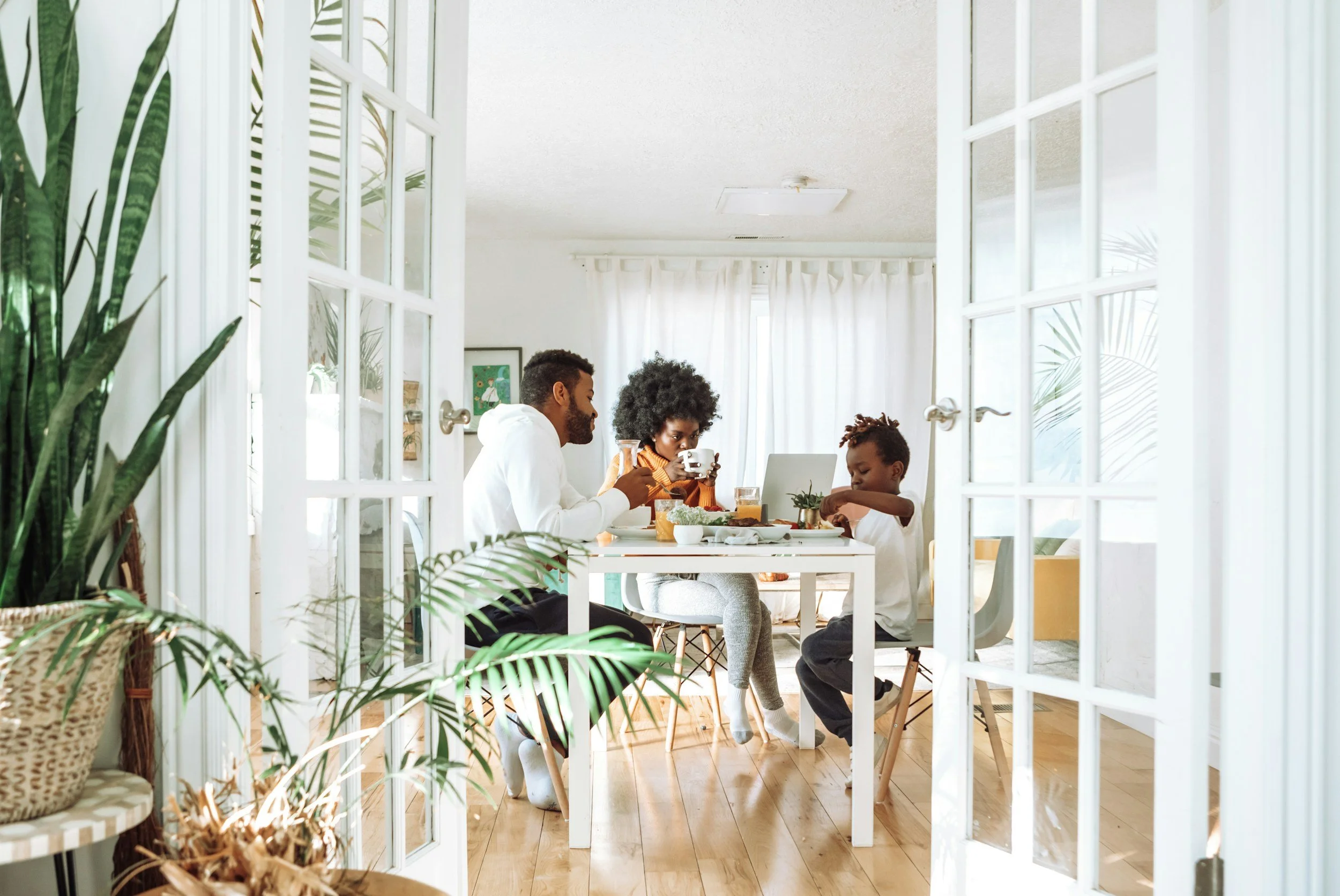 Family sitting at a dining table, having breakfast in a bright, airy room with white curtains, plants, and wooden flooring.