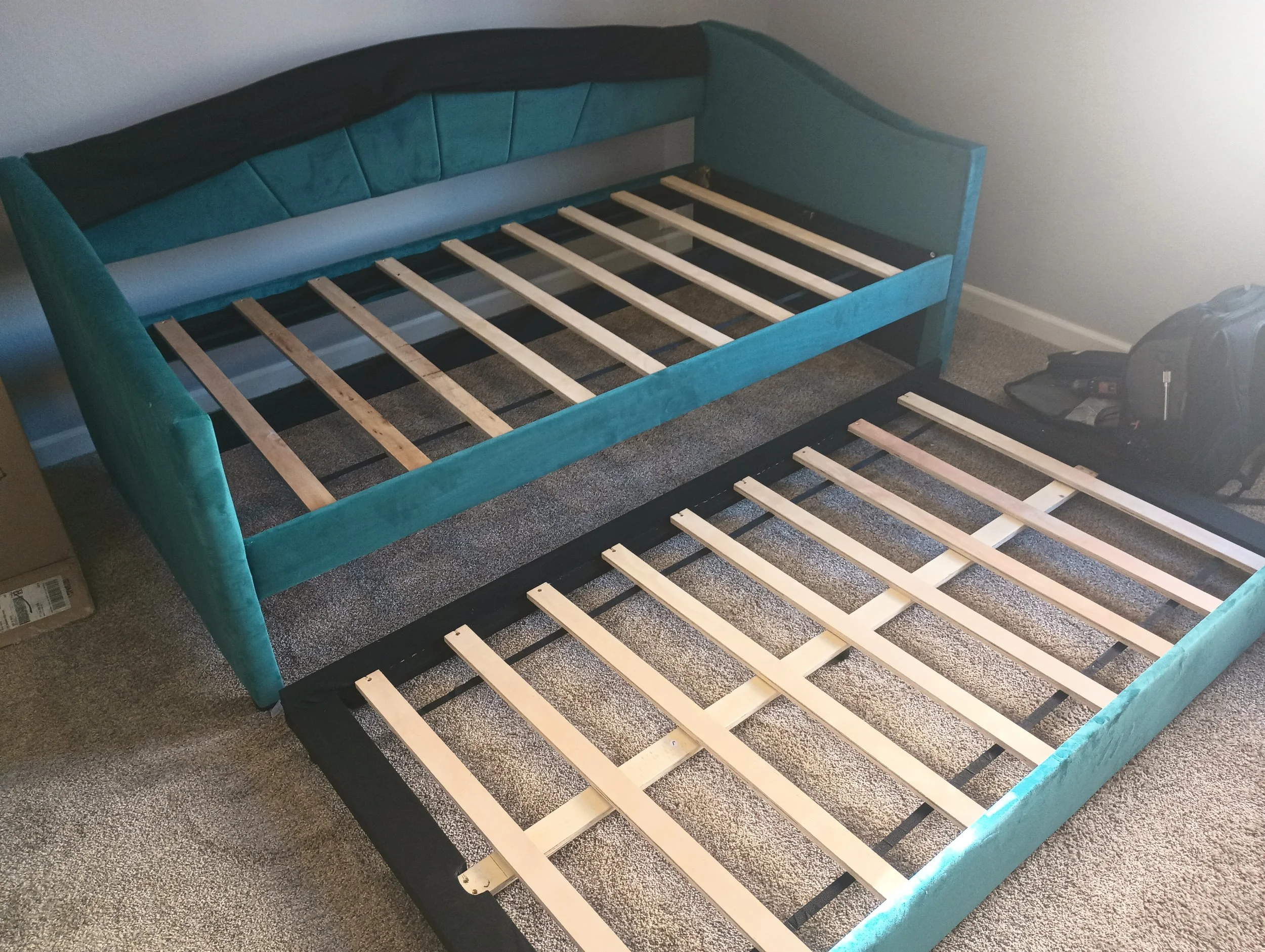 Two teal upholstered beds without mattresses, with wooden bed slats, placed on a carpeted floor in a room with plain walls. A backpack and a toolbox are on the floor next to one bed.