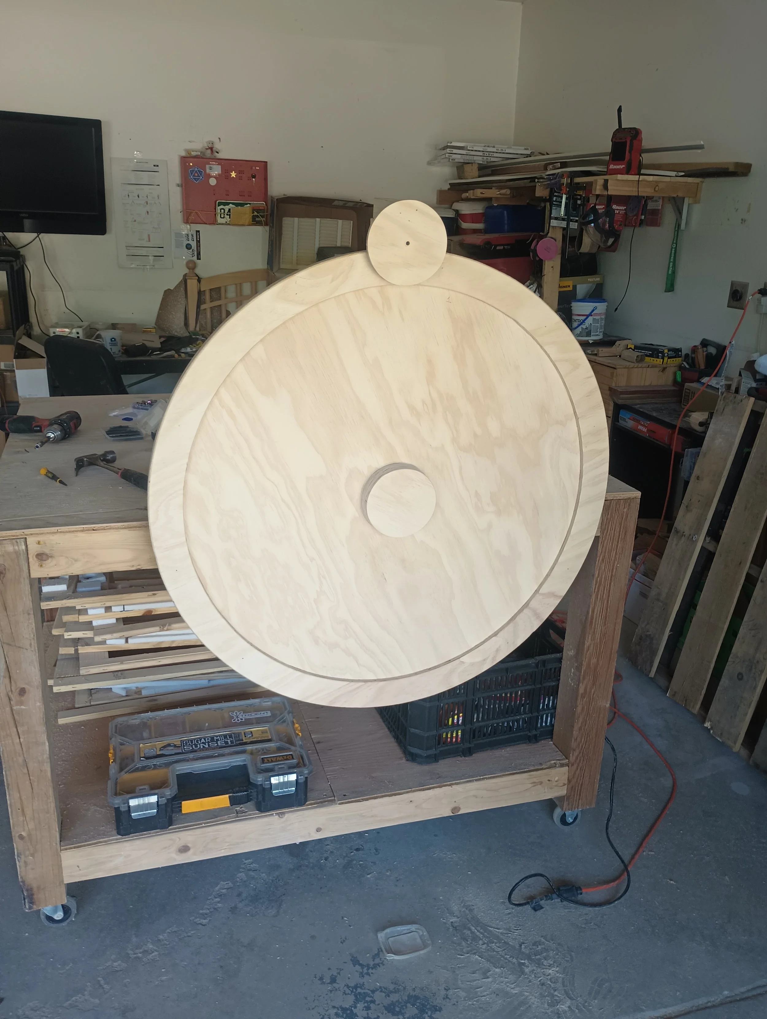 A large circular wooden structure with smaller wooden circles attached on top and in the center, placed on a workbench in a woodworking workshop.