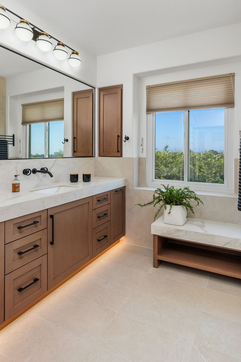 Modern bathroom with wooden cabinets, white marble countertop, under-sink lighting, and a large window with a view of greenery. A potted plant sits on a marble bench.