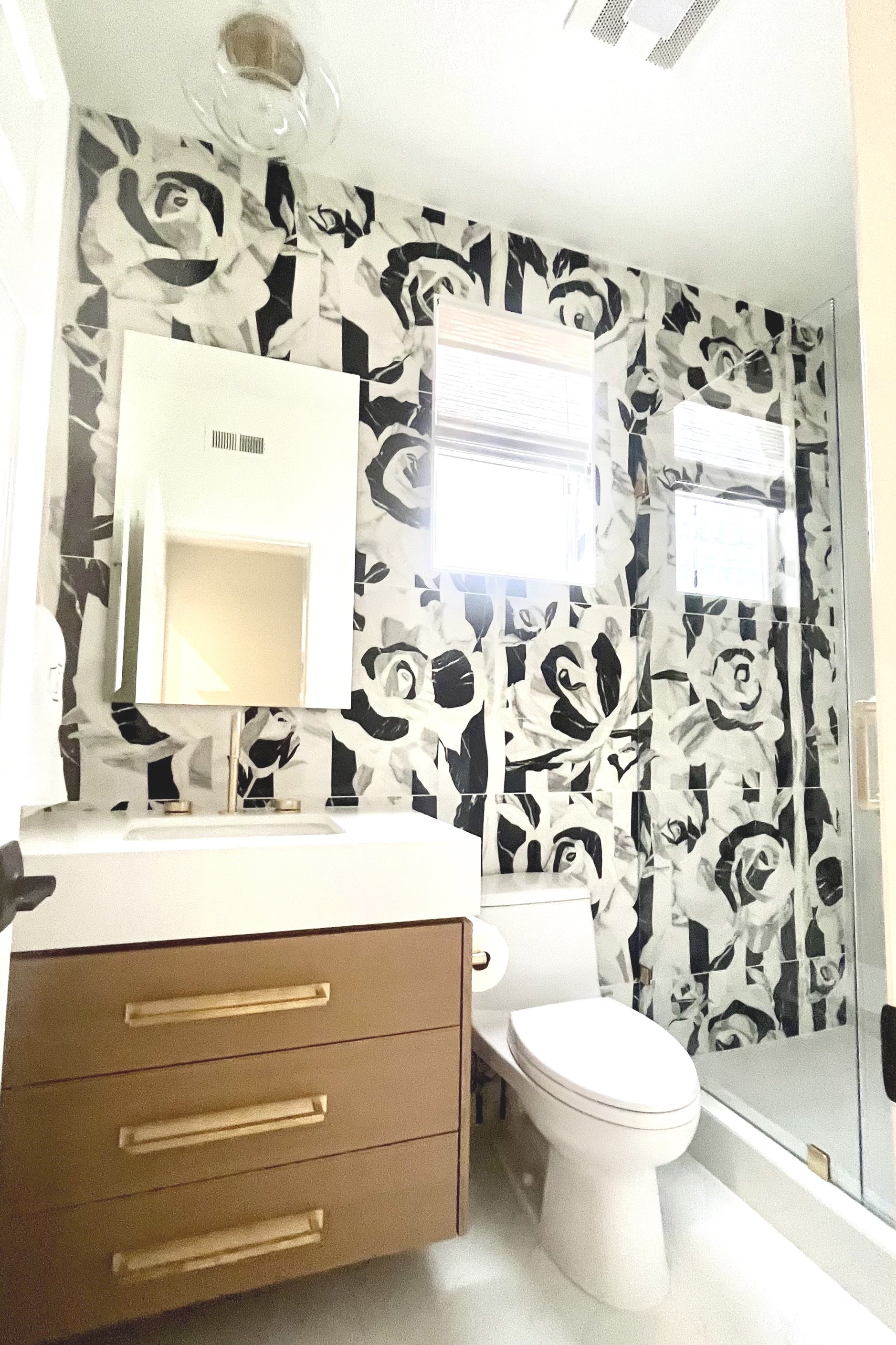 Bathroom with floral wallpaper, wood vanity with gold handles, white sink, toilet, mirror, and glass shower enclosure.