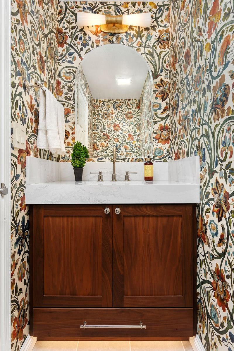 Small bathroom with floral wallpaper, a wooden vanity with a white countertop, a sink, a mirror, and a wall-mounted light fixture.