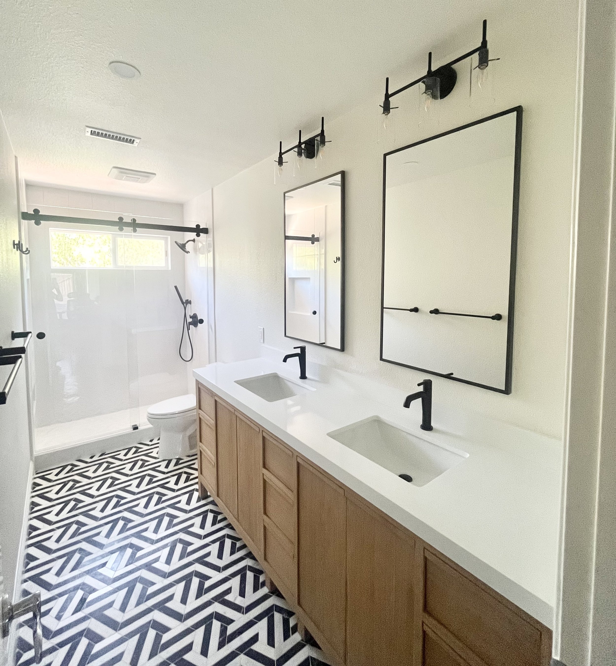 Modern bathroom with double sink vanity and black fixtures, geometric tile flooring, large mirrors, wall sconces, and a shower with sliding glass doors.