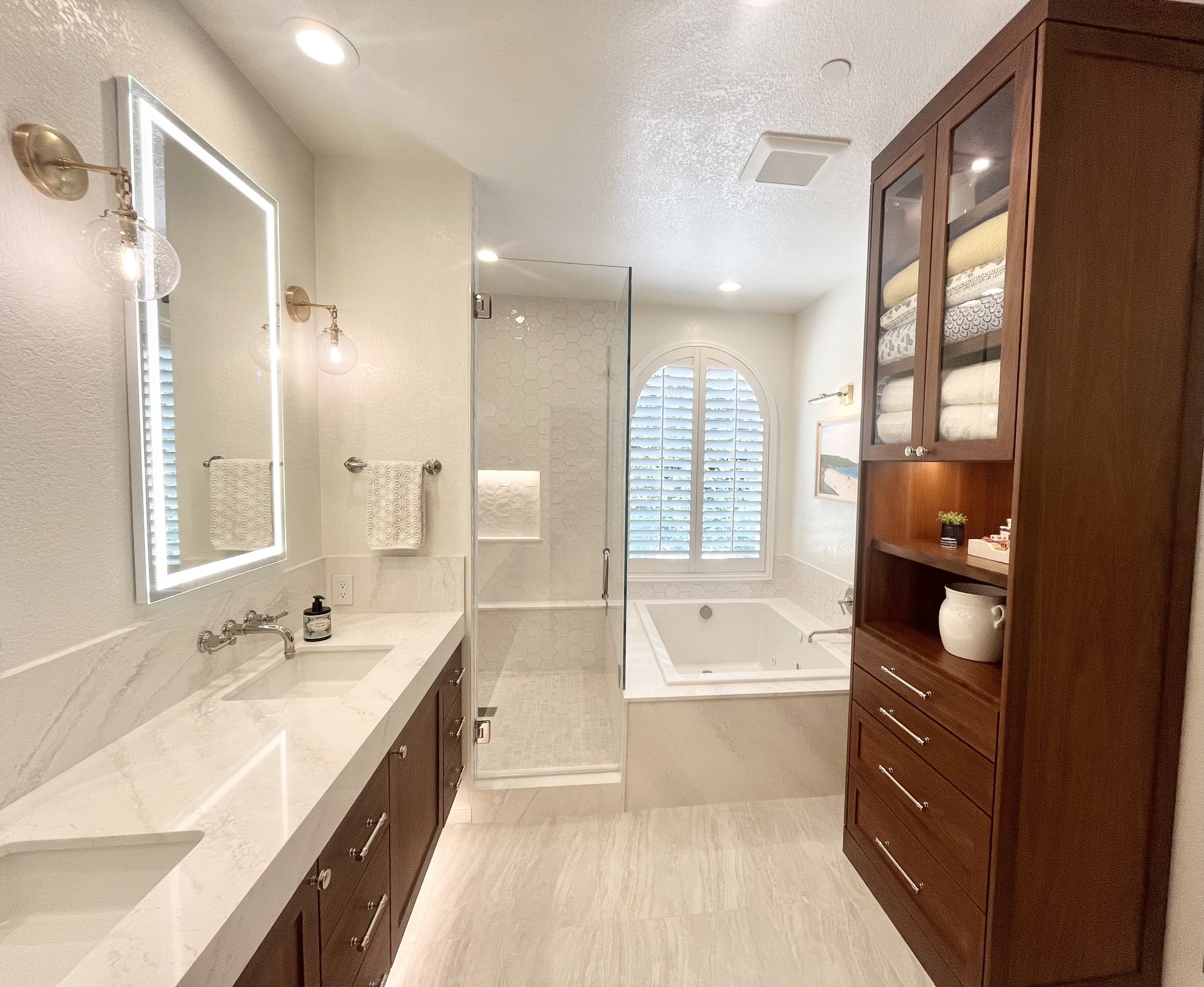 Modern bathroom with double sinks, glass shower, and built-in bathtub. Features include marble countertops, wooden cabinets, and a backlit mirror.