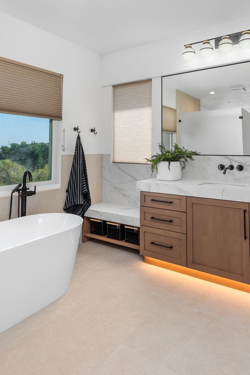 Modern bathroom with a white freestanding bathtub, wooden vanity with marble countertops, large mirror, under-cabinet lighting, potted plant, and striped towel. Beige tiles and window with blinds.