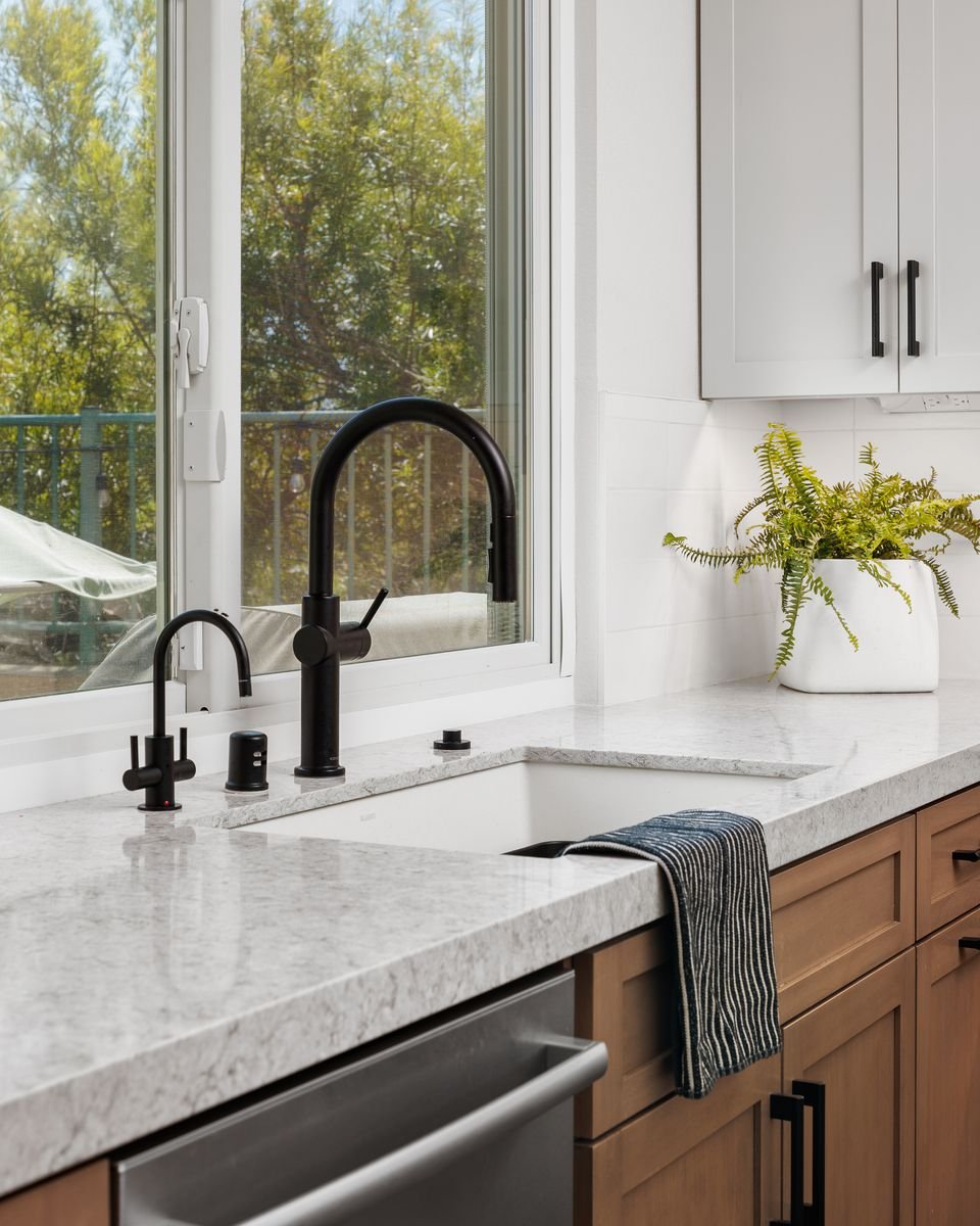 Modern kitchen sink with black faucet, soap dispenser, and a striped towel on a marble countertop. Wooden cabinets are below, with a potted plant beside a window.