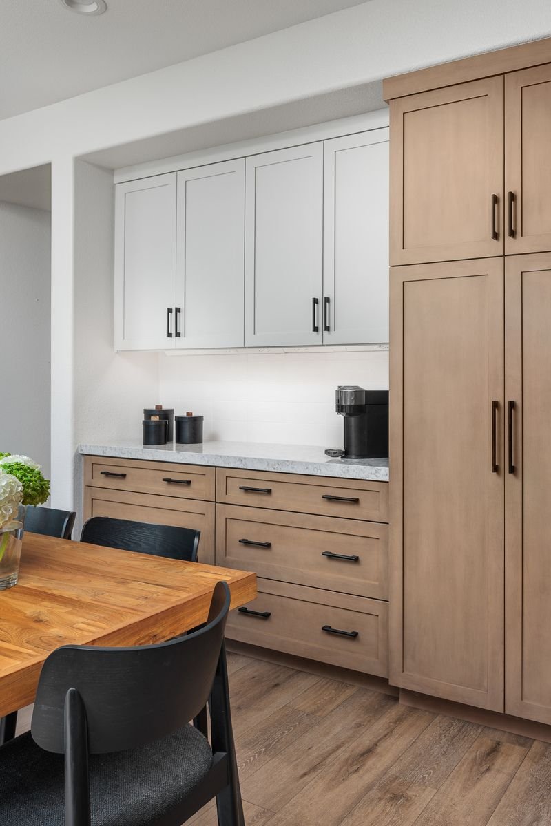 Modern kitchen with white and wood cabinets, black handles, gray countertop, coffee maker, wooden dining table, and black chairs.