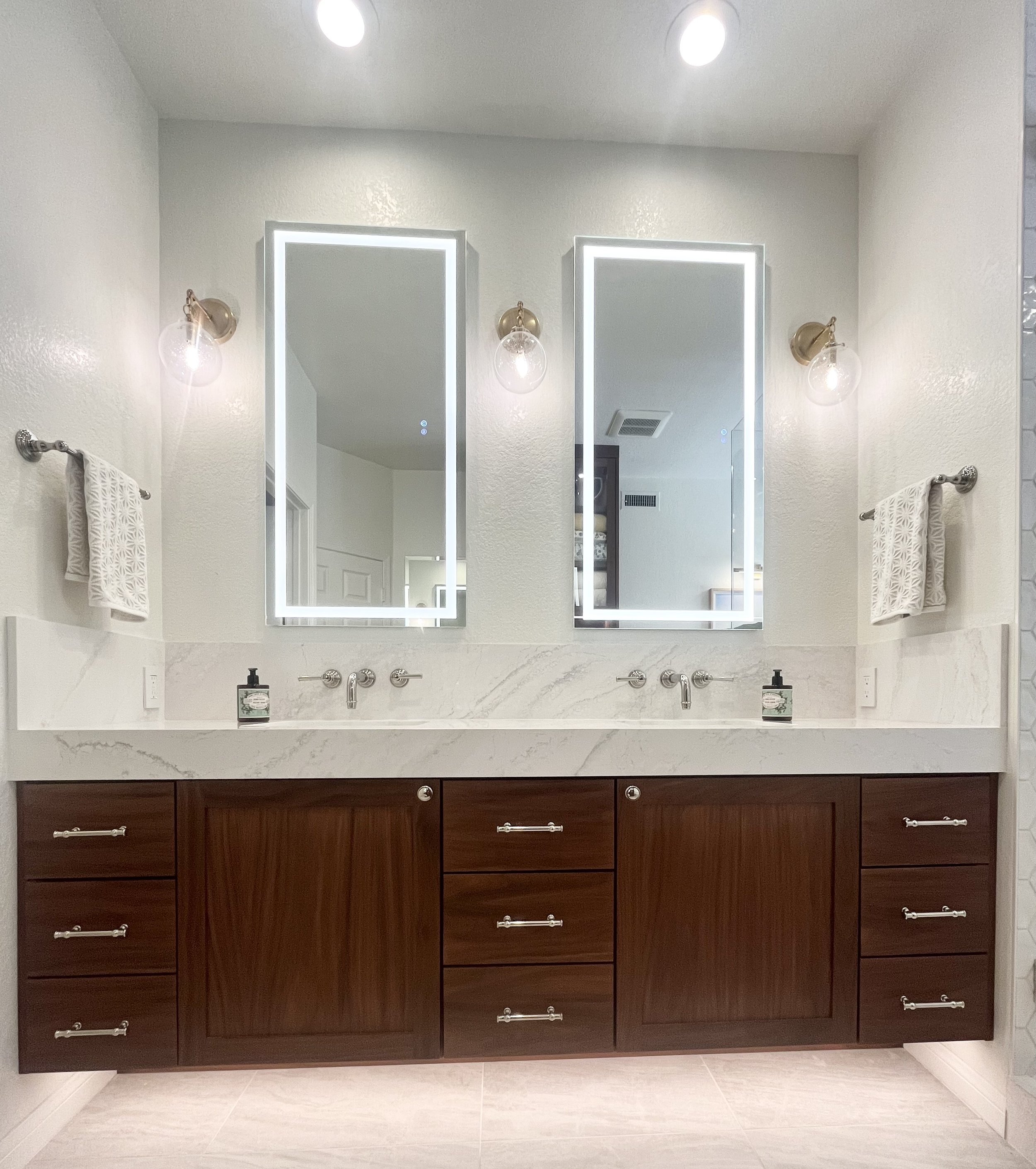 Modern bathroom vanity with two illuminated mirrors, dark wood cabinets, white marble countertop, two wall-mounted faucets, decorative light fixtures, and towels.