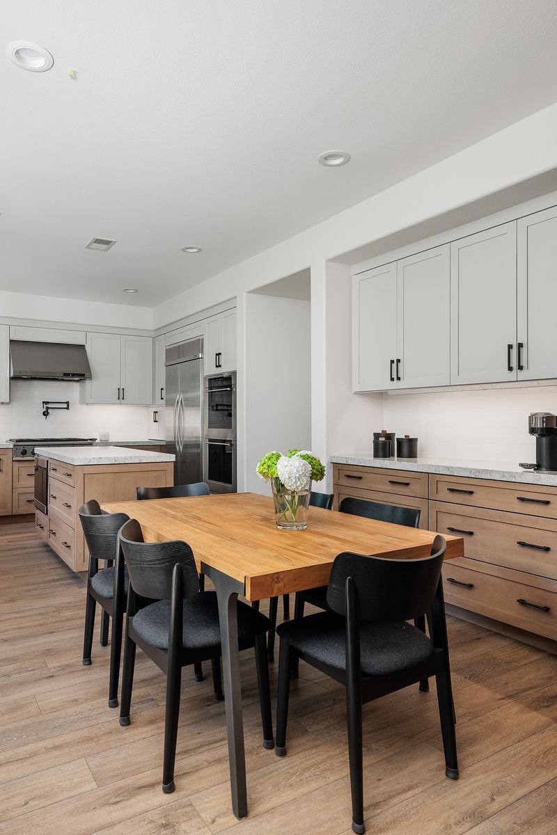 Modern kitchen with wooden dining table, black chairs, light gray cabinets, stainless steel appliances, and a vase with flowers.