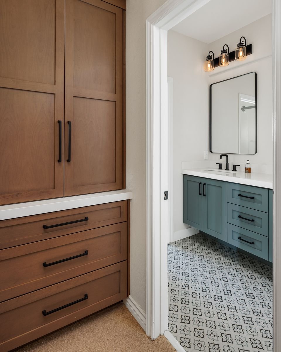 Interior with wooden cabinets and a view into a bathroom with blue vanity, patterned tile floor, and black light fixtures above a mirror.