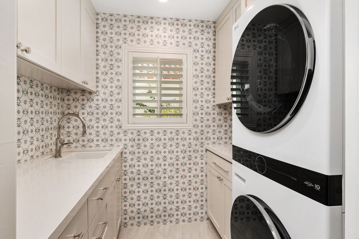 Modern laundry room with patterned tile backsplash, stacked washer and dryer, sink, white cabinets, and a window with shutters.