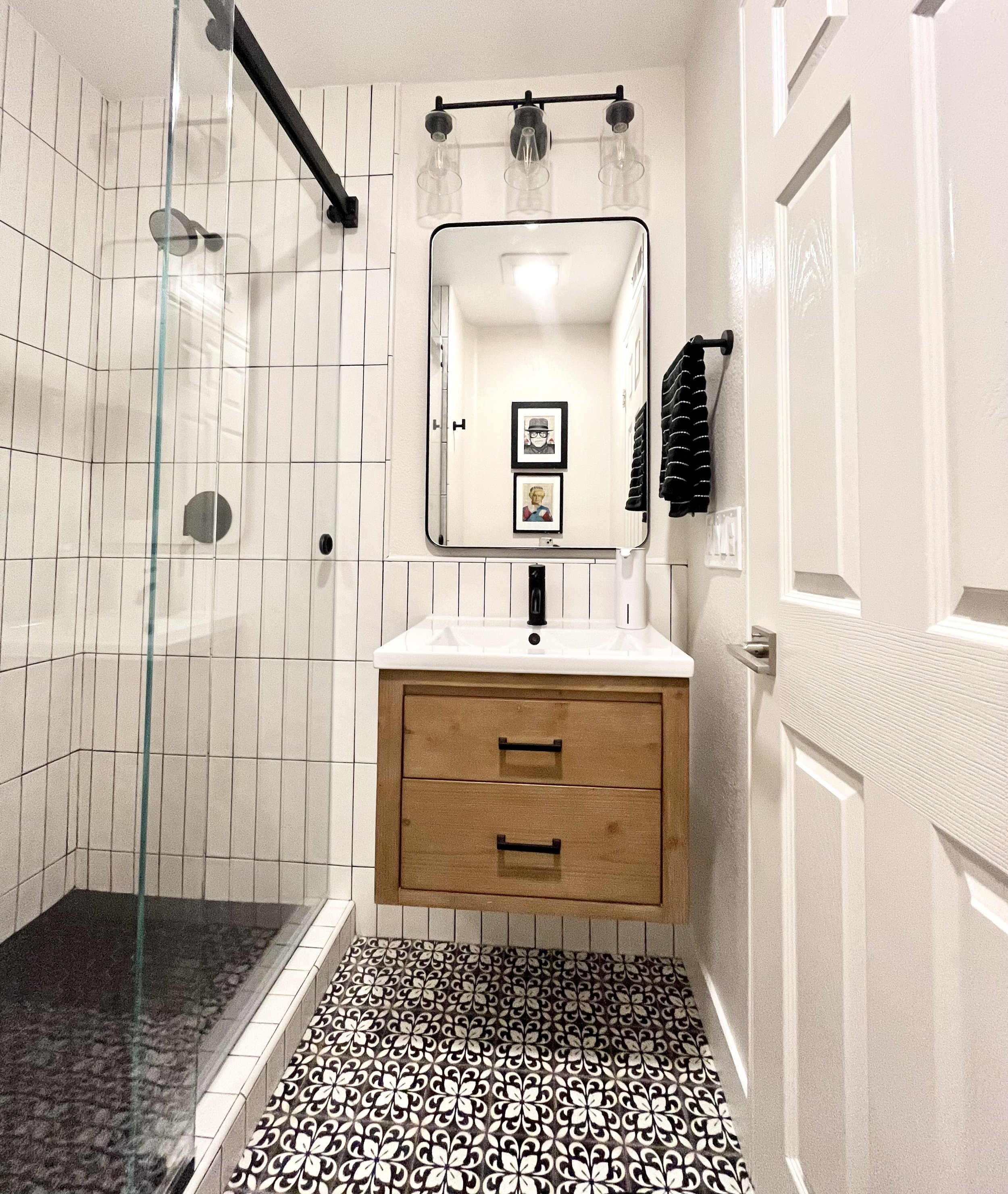 Modern bathroom with a glass shower, floating wooden vanity, geometric tile floor, white tiled walls, black fixtures, mirror, and wall-mounted light fixture.