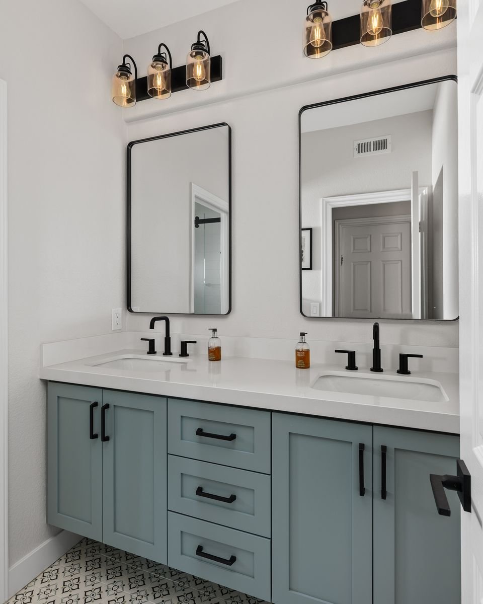 Modern bathroom with twin sinks, teal cabinetry, black fixtures, two mirrors, and industrial-style light fixtures.