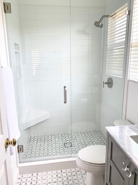 Modern bathroom with glass-enclosed shower, white tiled walls, patterned floor tiles, toilet, and vanity with a marble countertop.