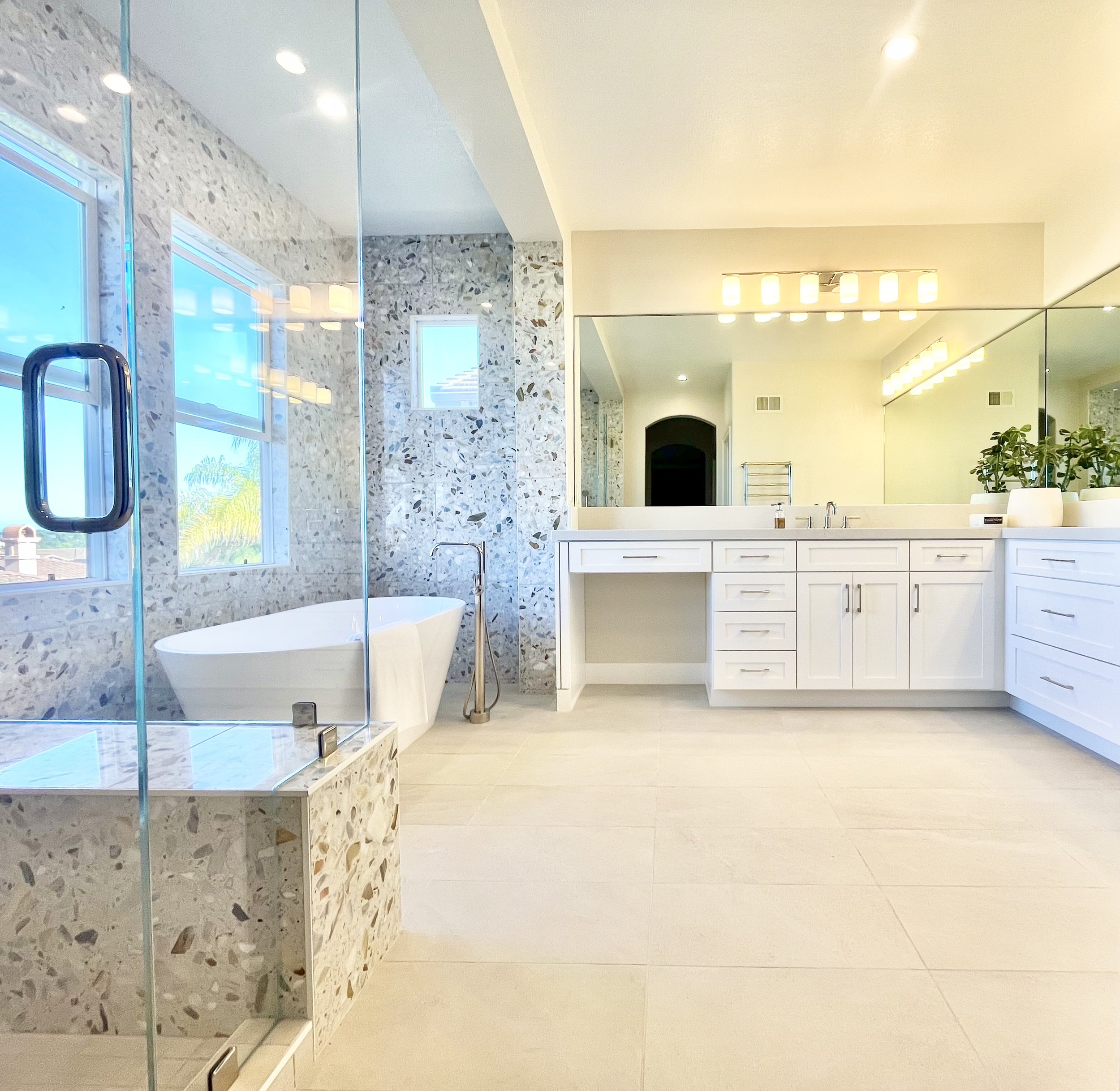 Modern white bathroom with large mirror, double vanity with white cabinets, freestanding bathtub, glass shower, terrazzo tiles, and potted plants.