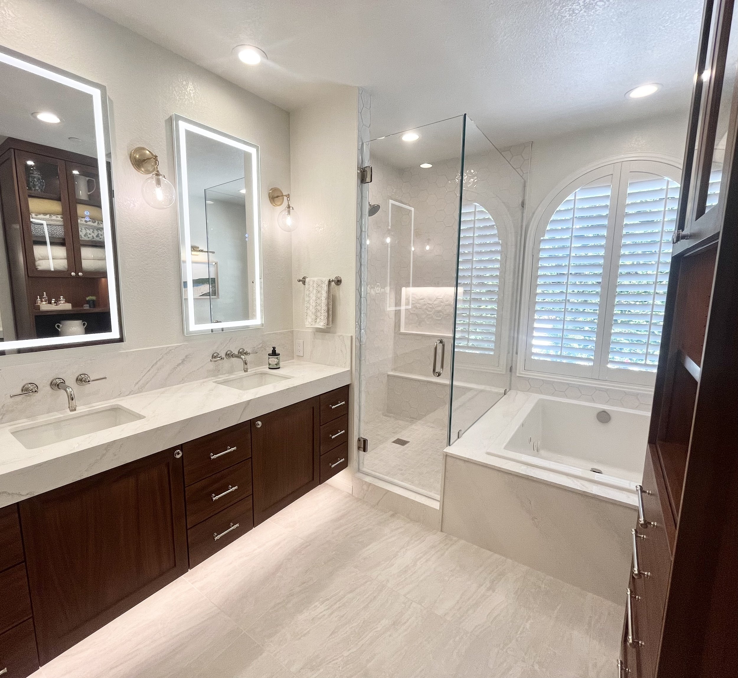 Modern bathroom with double sink vanity, glass shower, and bathtub. Features white countertops, wood cabinets, and large windows with shutters.