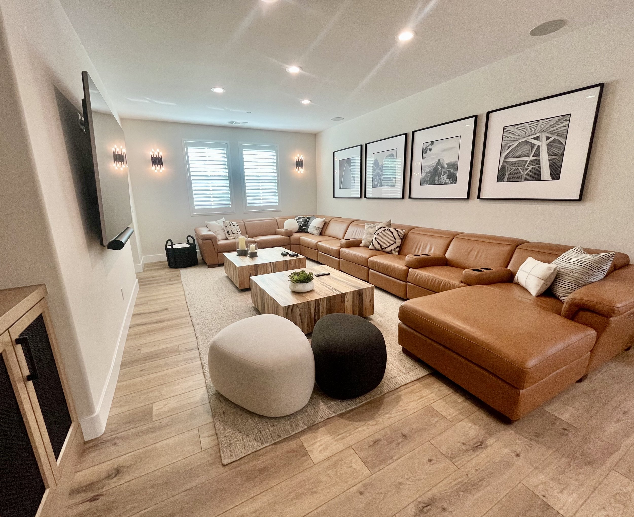 Modern living room with a large brown leather sectional sofa, wooden coffee table, two poufs, wall-mounted TV, framed monochrome art, and decorative lighting.