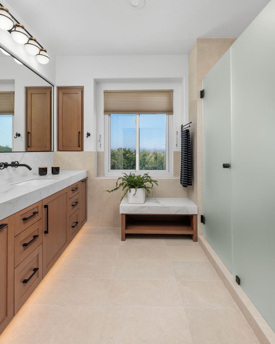Modern bathroom interior with wooden cabinets and white countertop, large mirror, a plant, window with blinds, black towel on a rod, and glass shower door.