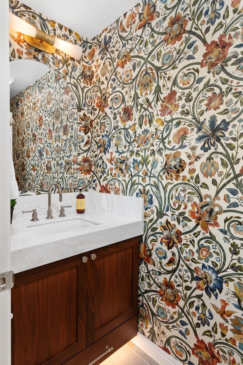 Bathroom with floral wallpaper, white marble sink, wooden vanity, and wall-mounted light fixture.