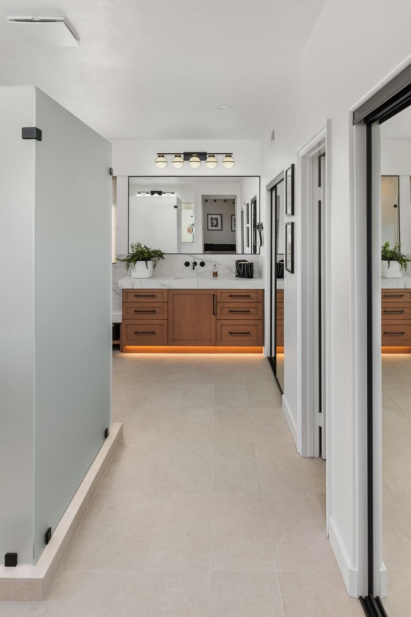 Modern bathroom with wooden vanity, under-cabinet lighting, dual sinks, and wall mirror; frosted glass shower on left and tiled floor.