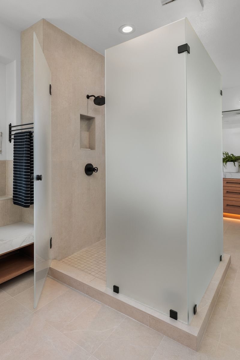 Modern bathroom with frosted glass shower enclosure, beige tiles, and a black towel rack with folded dark towel.