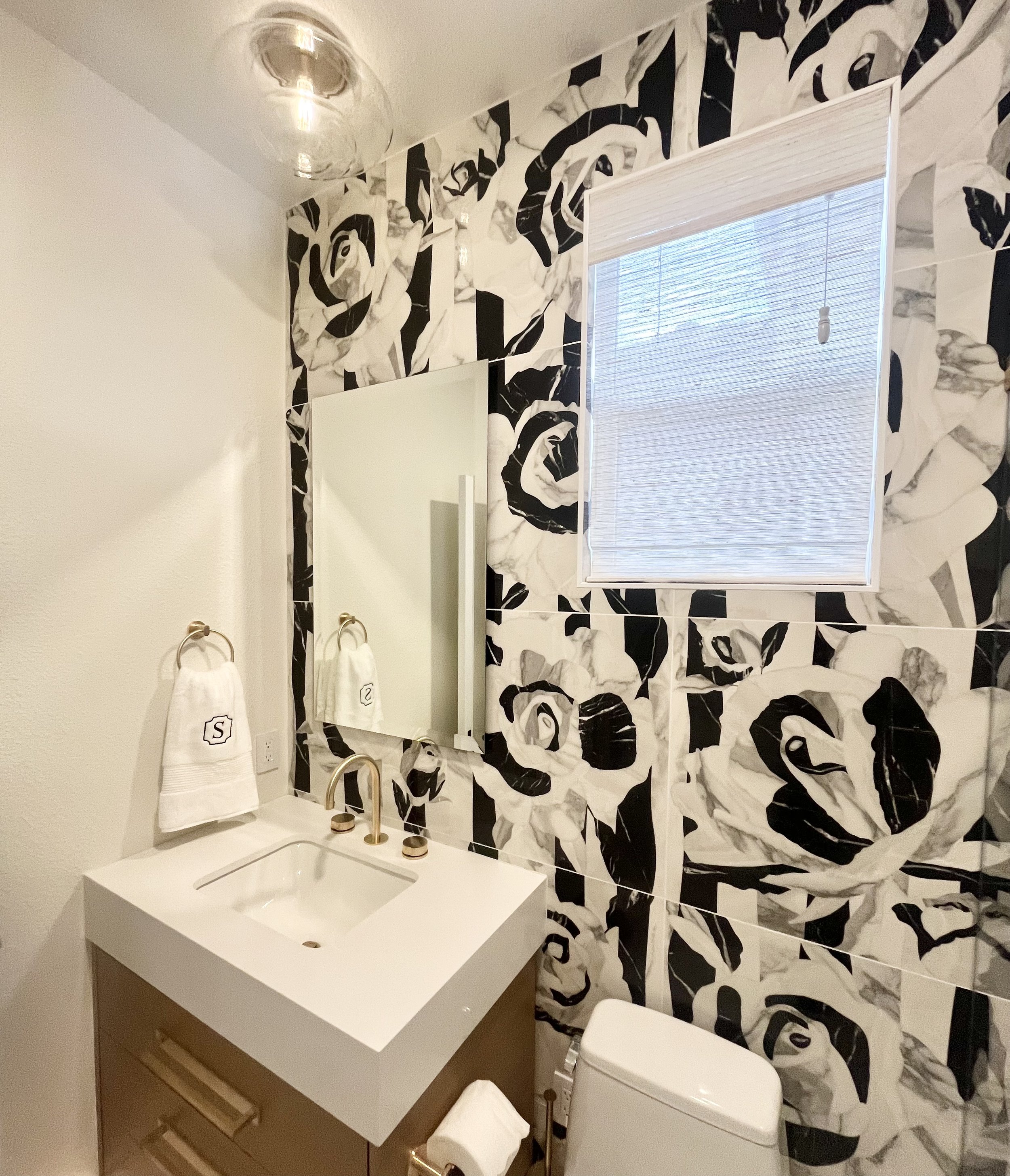 Bathroom with floral black and white wallpaper, modern vanity, square sink, gold fixtures, and a window with a blind partially down.
