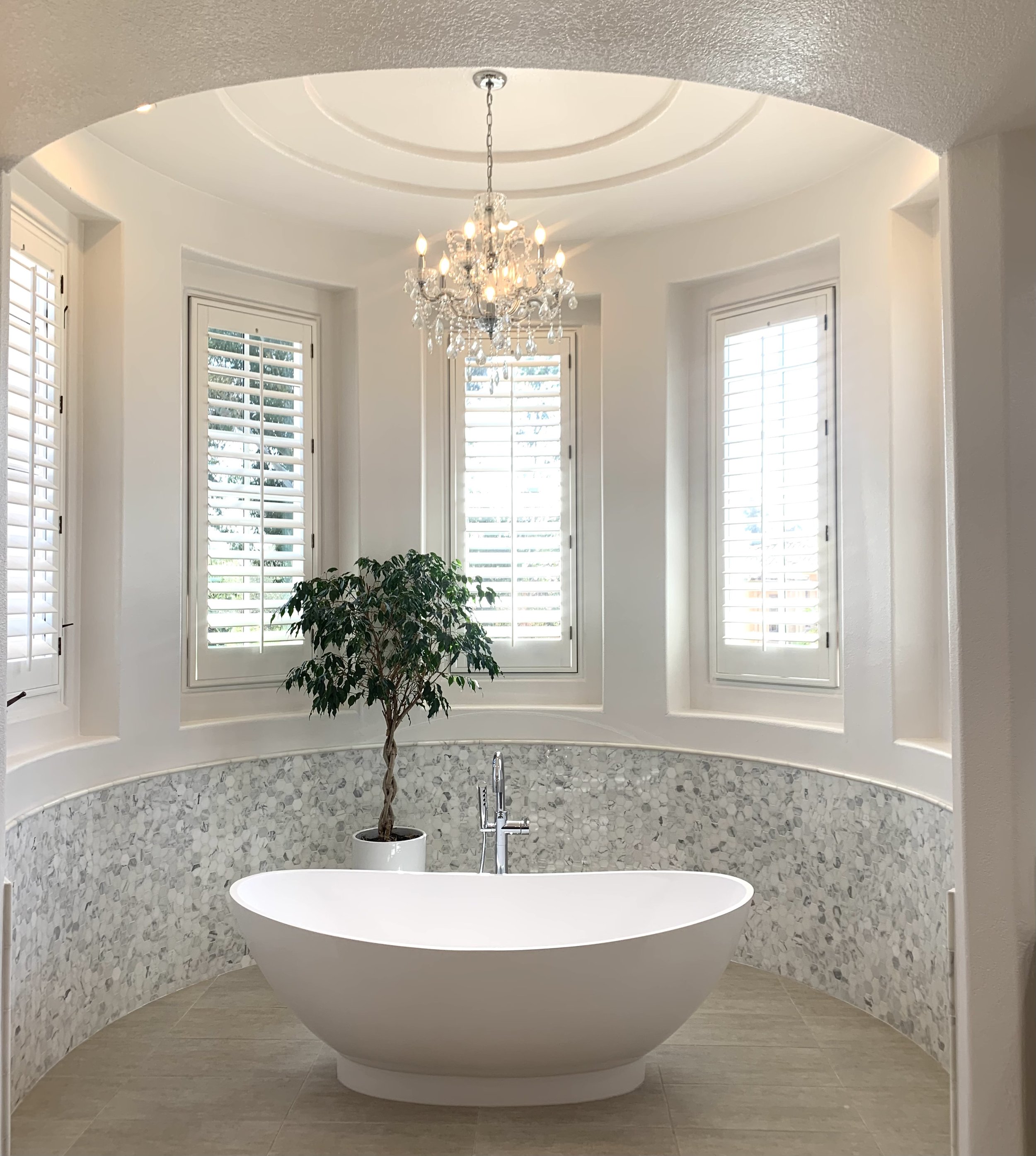 Luxury bathroom with white freestanding bathtub, crystal chandelier, potted plant, and large windows with shutters.