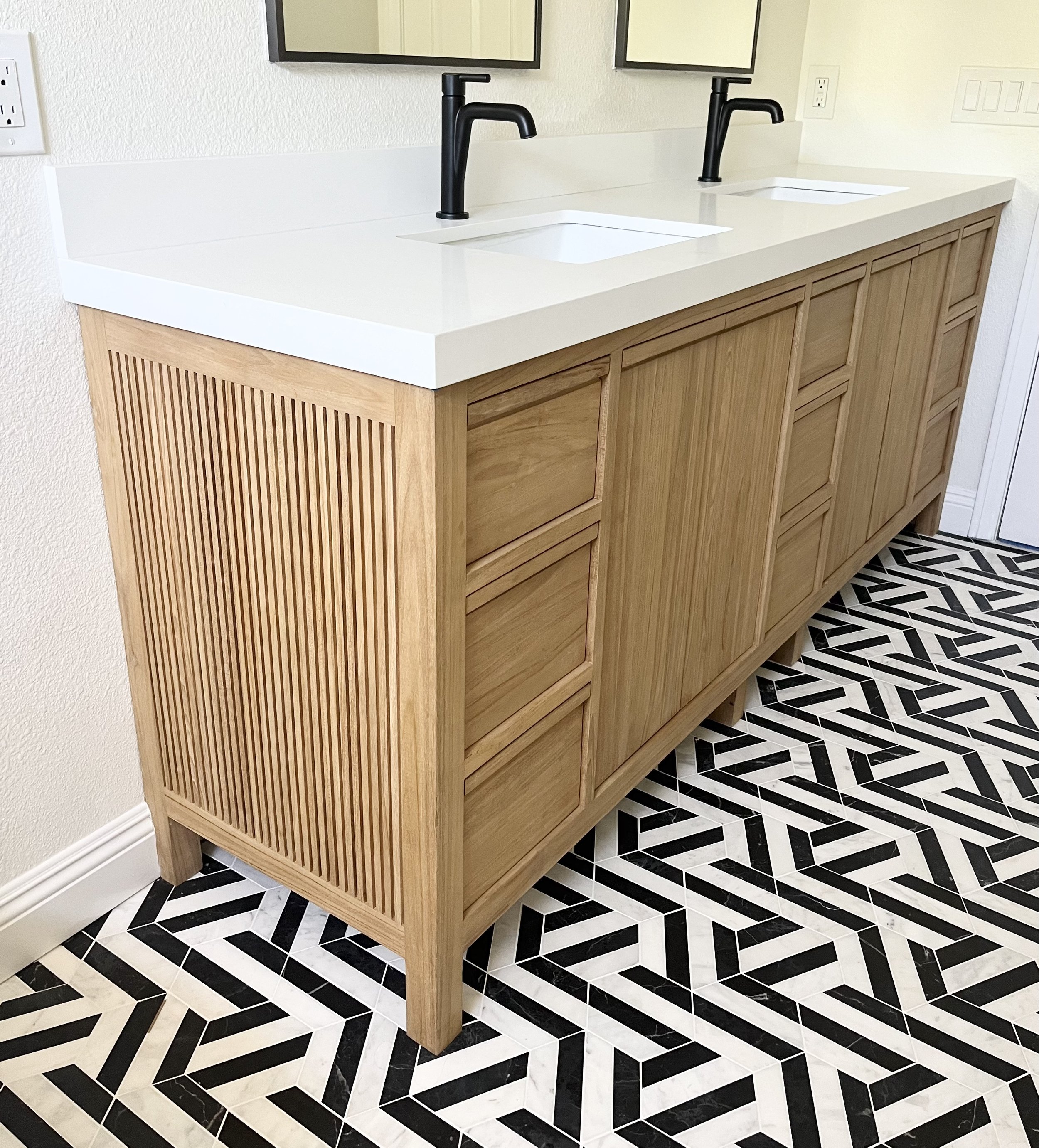 Double bathroom vanity with white countertops, black faucets, and a natural wood finish. The floor features a black and white geometric pattern.