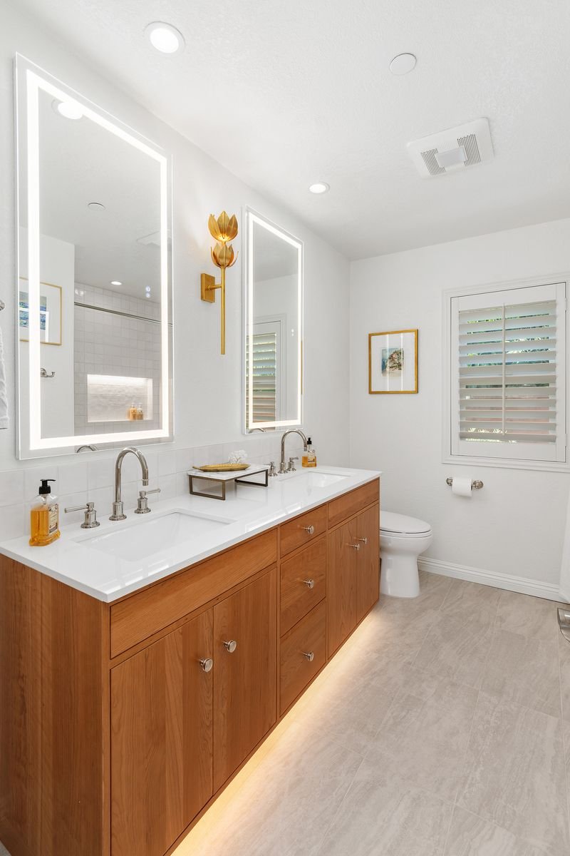 Modern bathroom with double vanity, illuminated mirrors, wooden cabinetry, toilet, and decorative wall art.