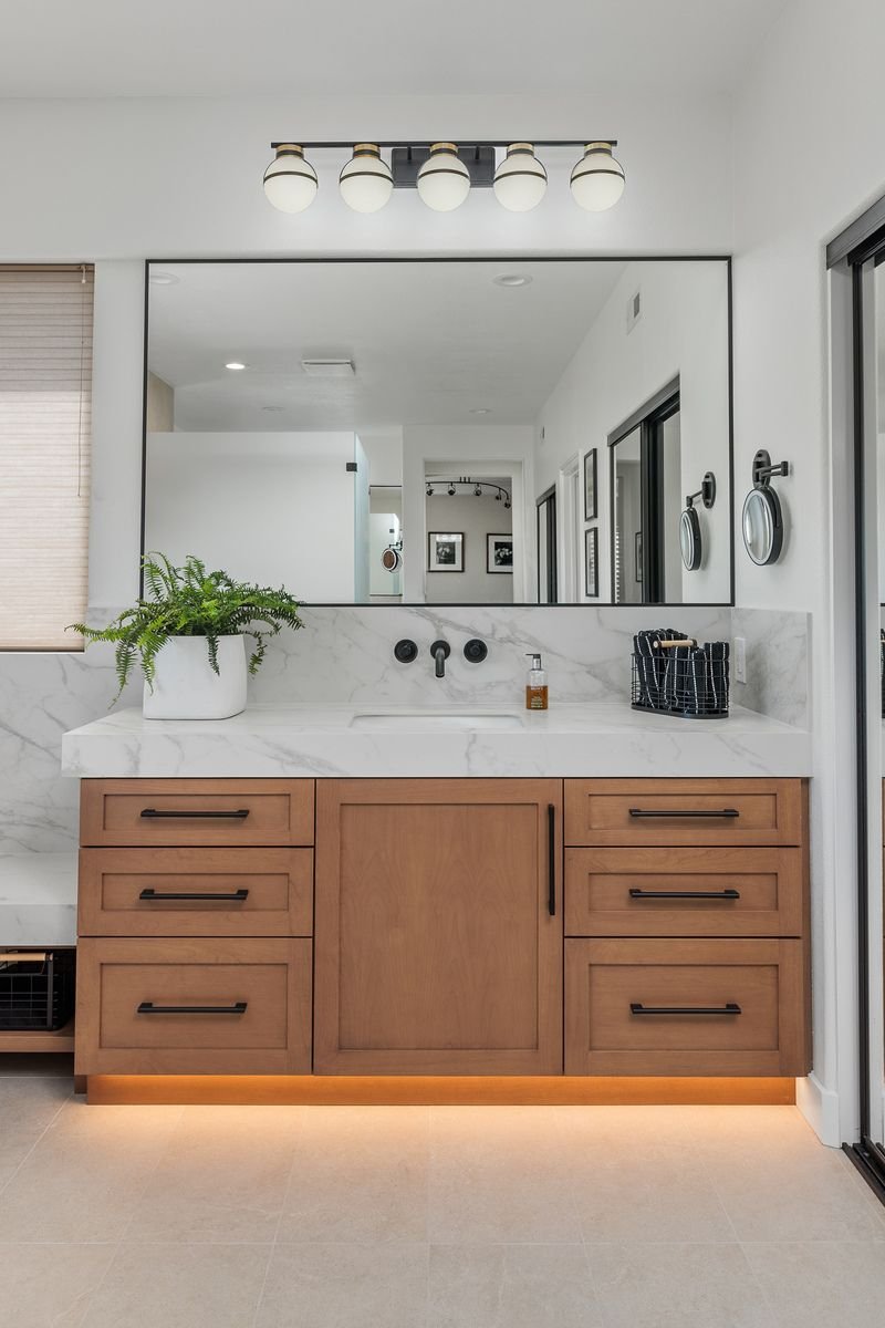 Modern bathroom vanity with a large mirror, wooden cabinets, and under-cabinet lighting. A countertop features a potted plant, soap dispenser, and decorative basket.