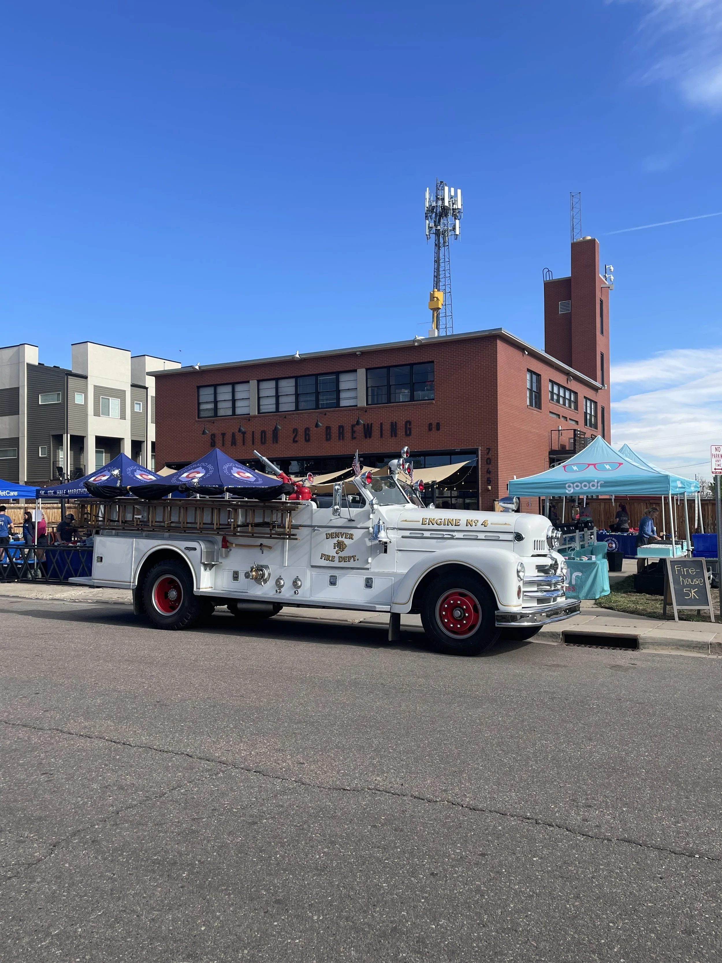 A vintage fire truck parked on a street at a local event with tents and people. The truck has the text 'Engine No. 4 Denver Fire Dept.' on the side. There are tents and a building in the background with the sign 'Station 26 Brewing'.