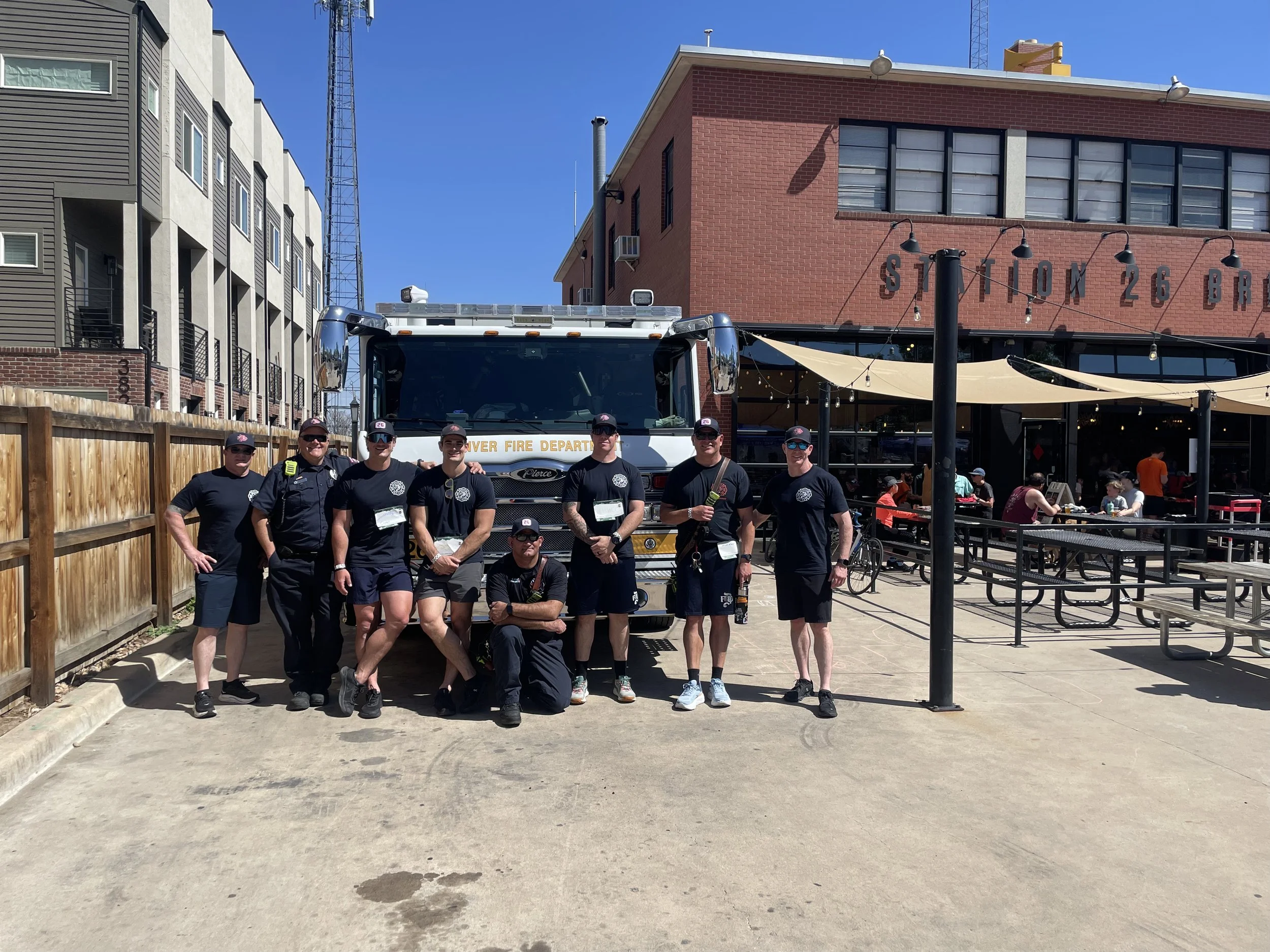 A group of six firefighters standing in front of a fire truck, smiling, in an outdoor urban area with buildings, a fence, and outdoor seating under a canopy.