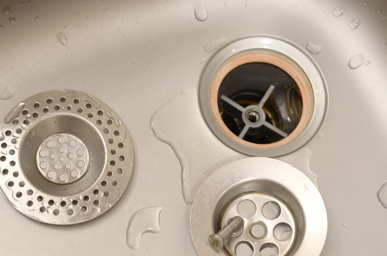 Close-up of a stainless steel kitchen sink drain with water droplets, showing two drain covers and the sink opening with an cross-shaped object inside.