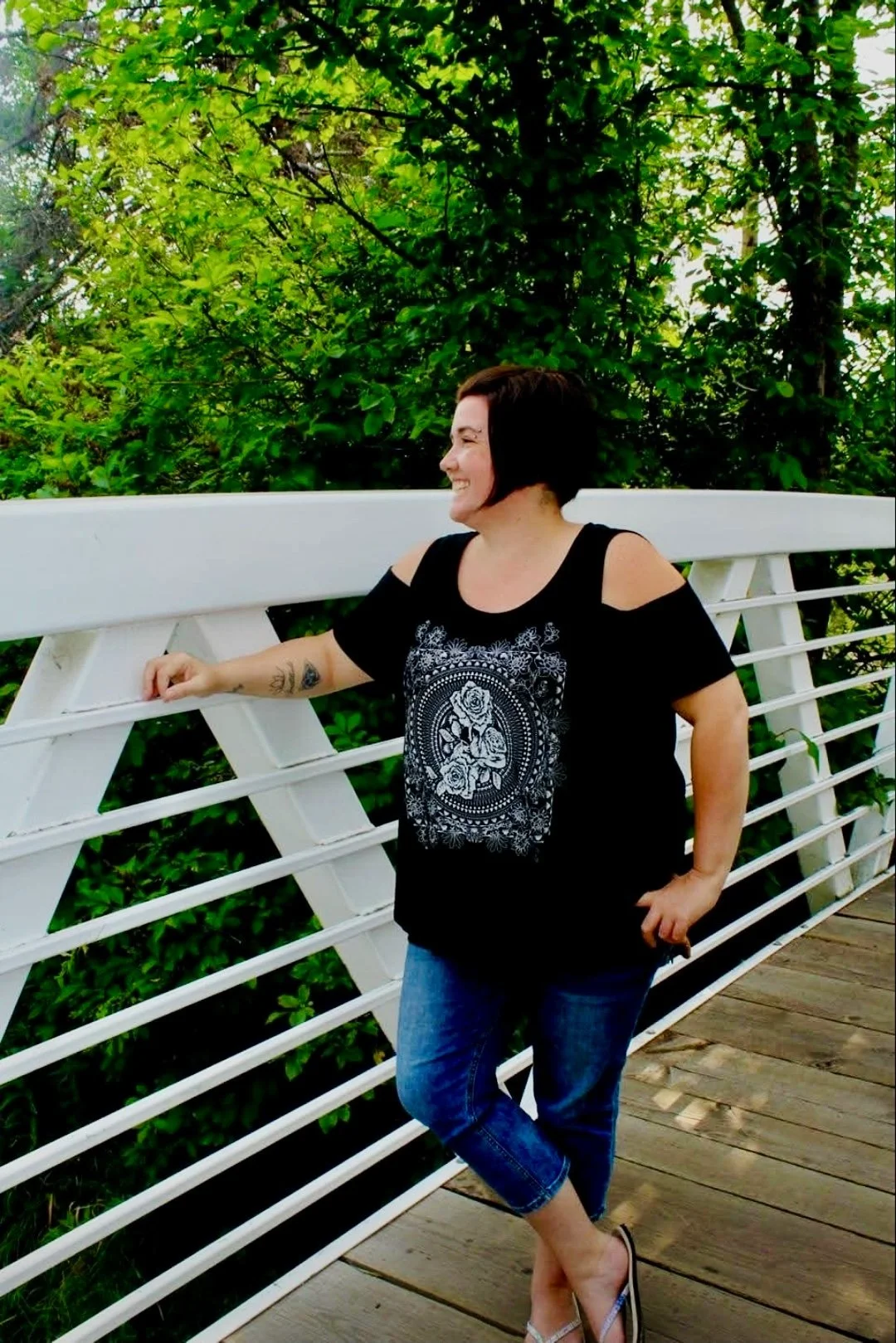Smiling woman with short dark hair, wearing a black top with cut-out shoulders and a white floral design, standing on a wooden bridge with white railing, surrounded by green trees.