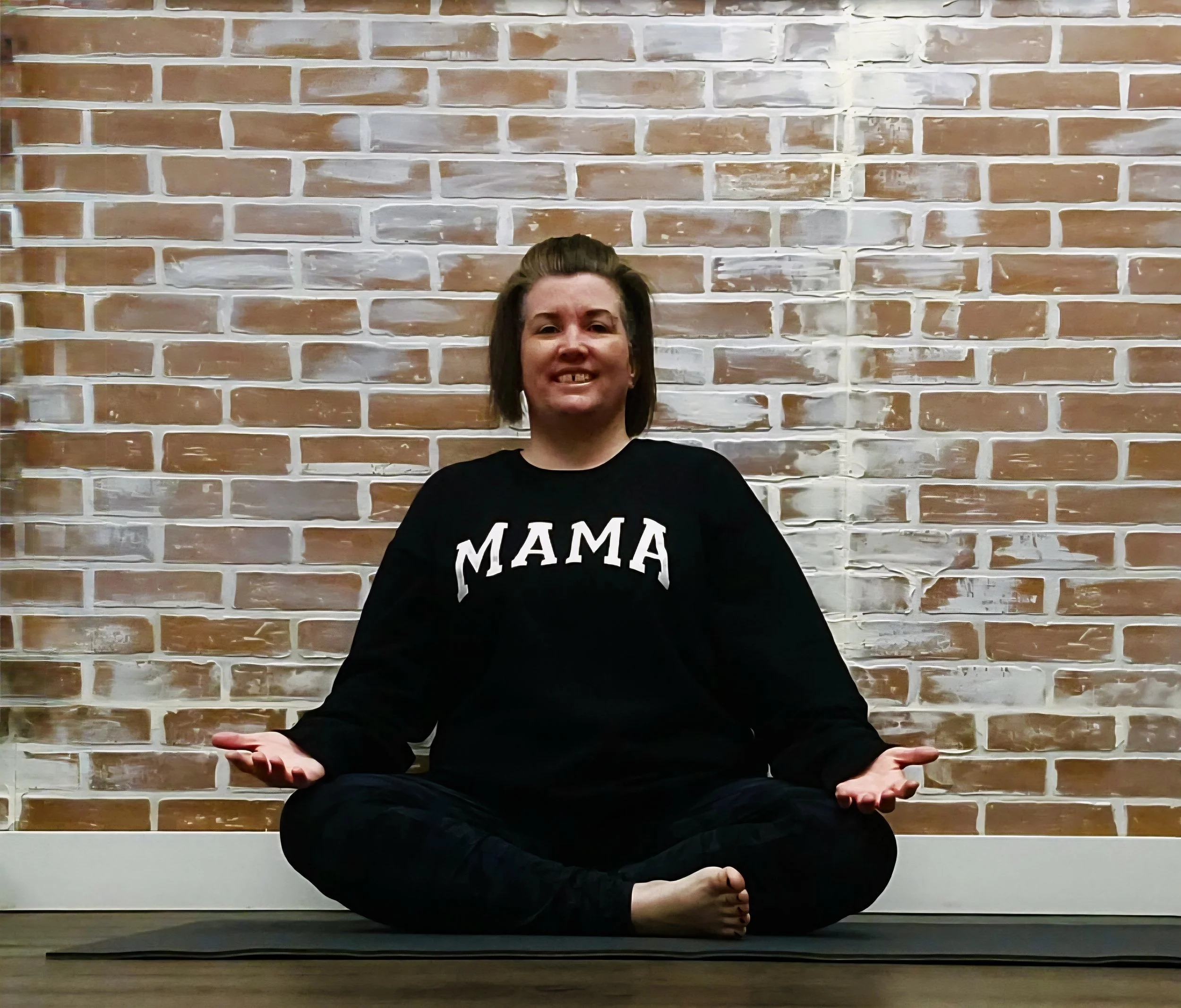 Woman in a black sweatshirt with 'MAMA' printed on it, sitting cross-legged on a yoga mat against a brick wall, practicing meditation or yoga.