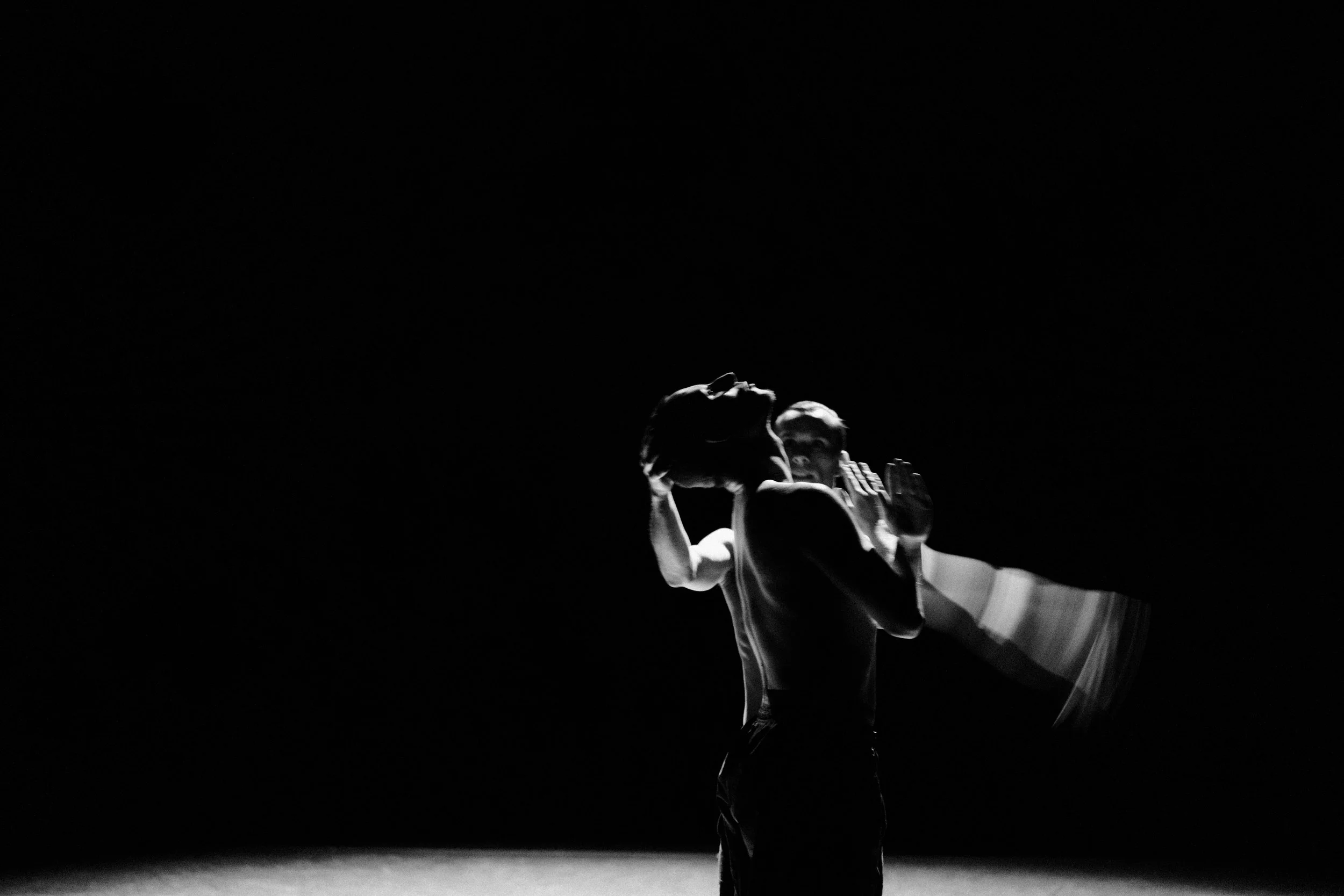 A person performing a dance move on stage with dramatic lighting, showing a mid-air pose with arms bent and head tilted back.