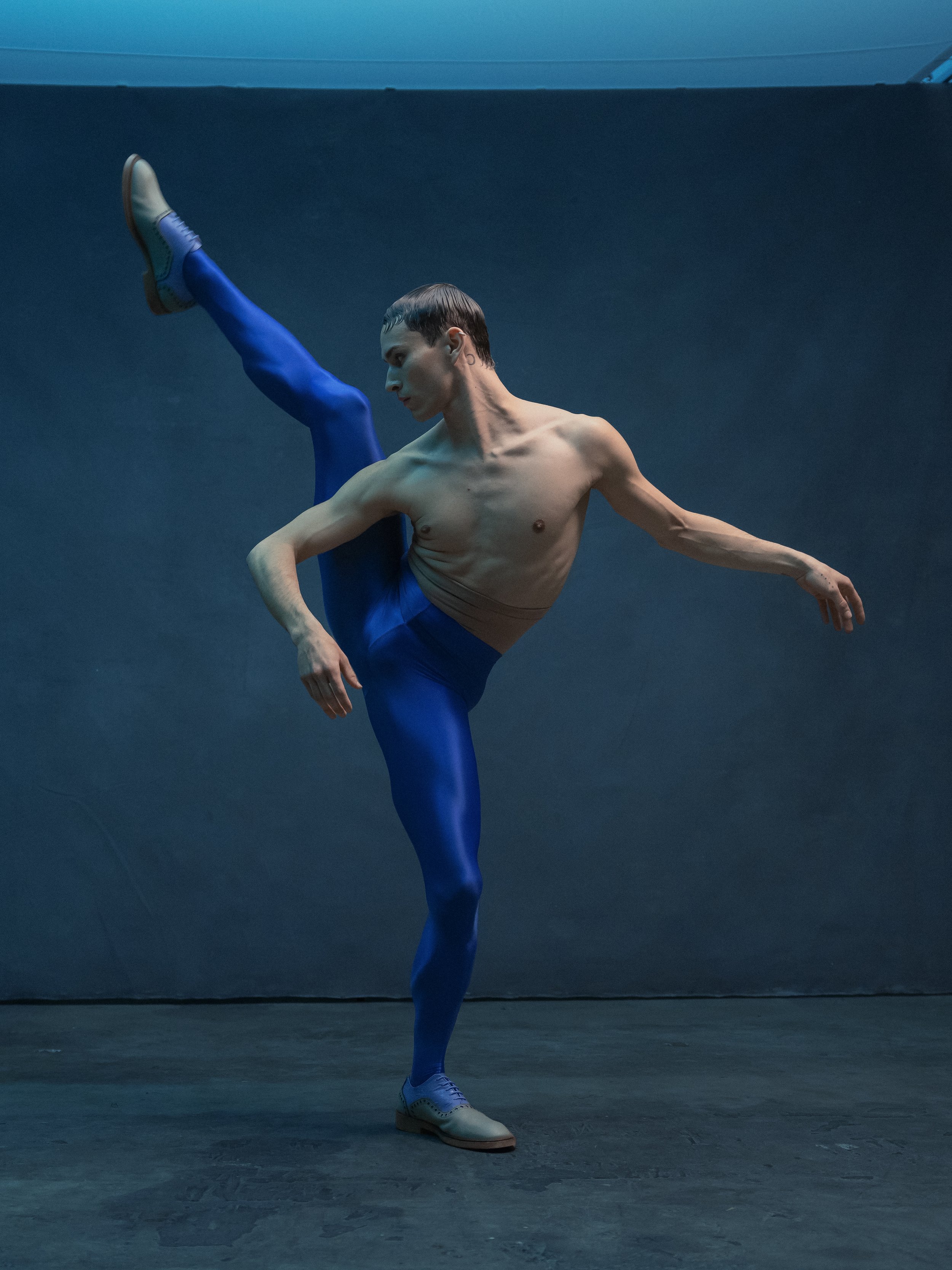 A male ballet dancer performing a high leg kick, wearing blue tights, beige shoes, and a shirtless torso, against a dark background.