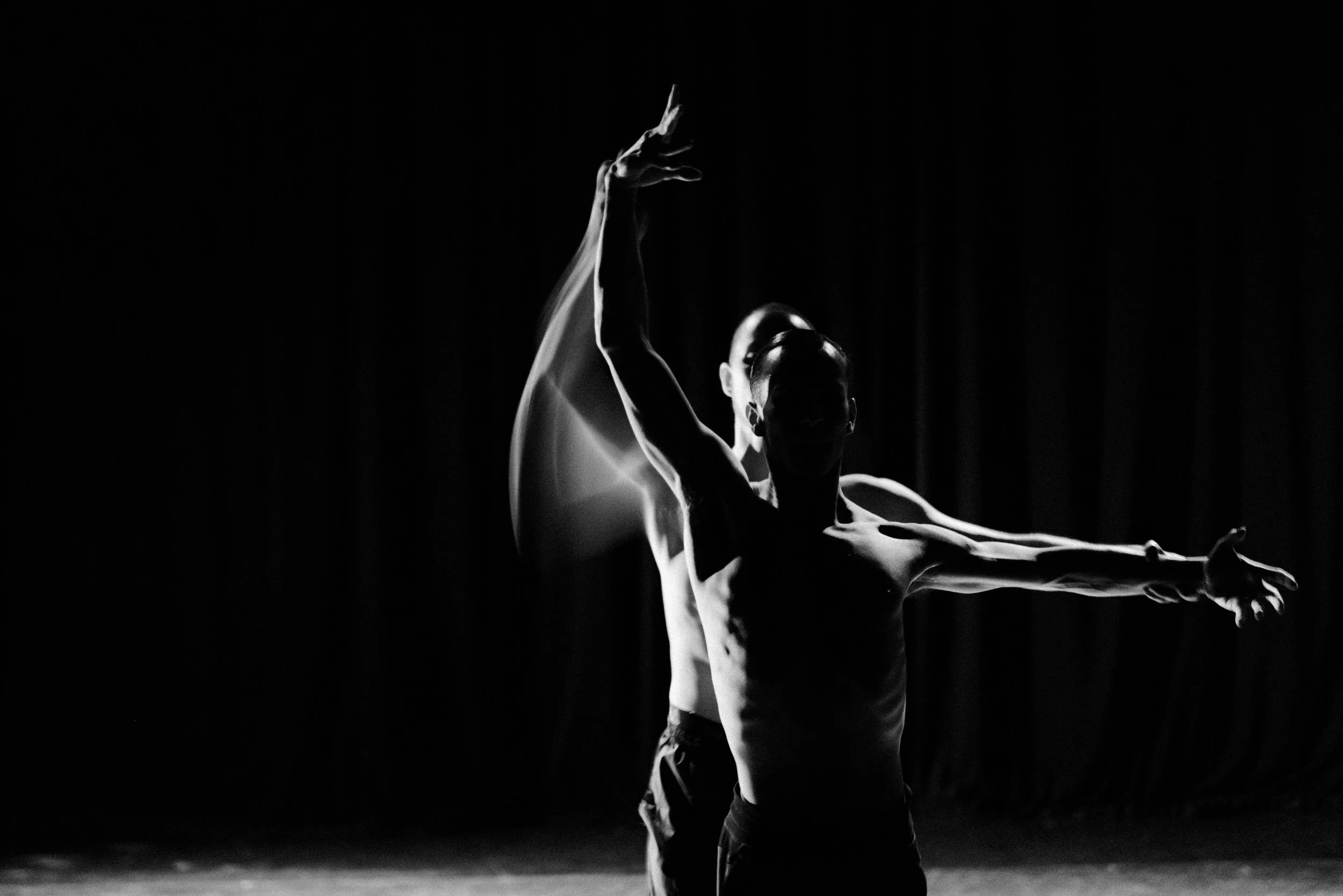 A dancer performs on stage, silhouetted against darkness, with one arm raised and head tilted back.