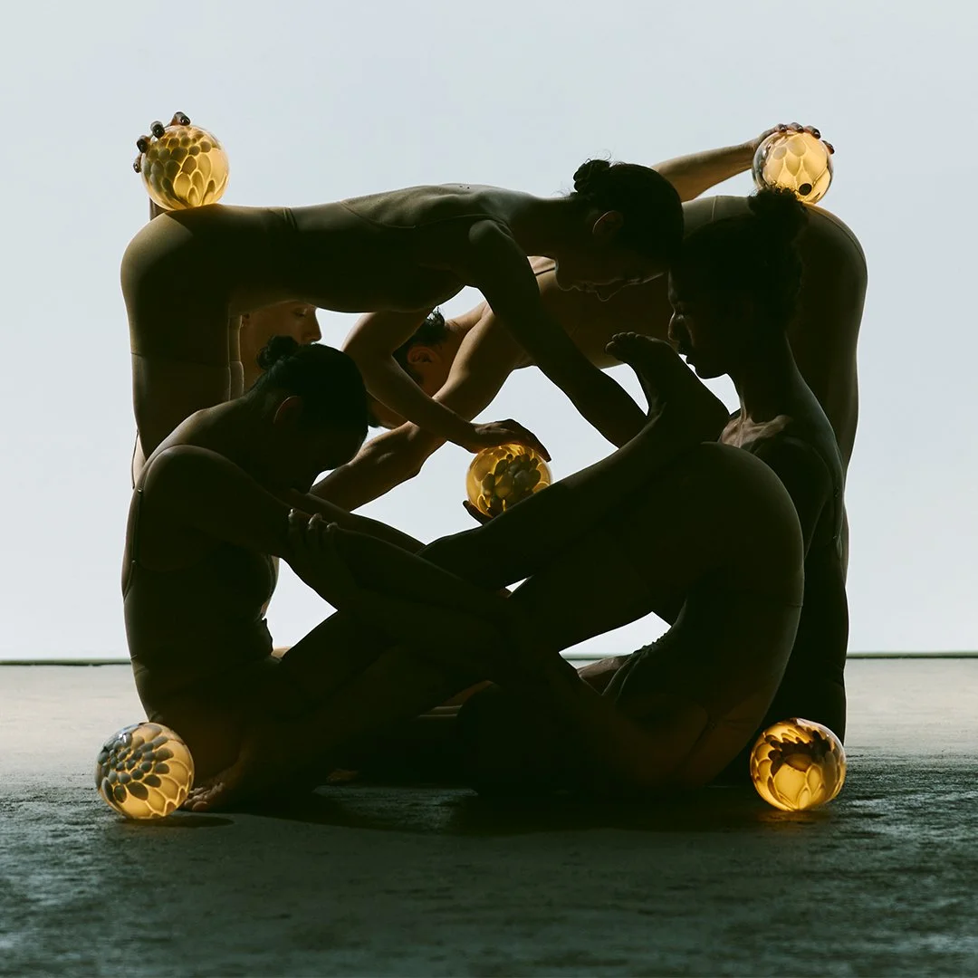 Silhouettes of five dancers in poses with illuminated orbs on their bodies and around them against a plain background.