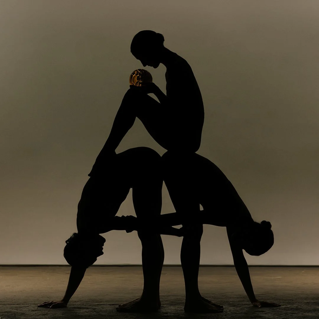 Silhouettes of three people performing acro yoga, with a small golden globe balanced on one person's head, against a neutral background.