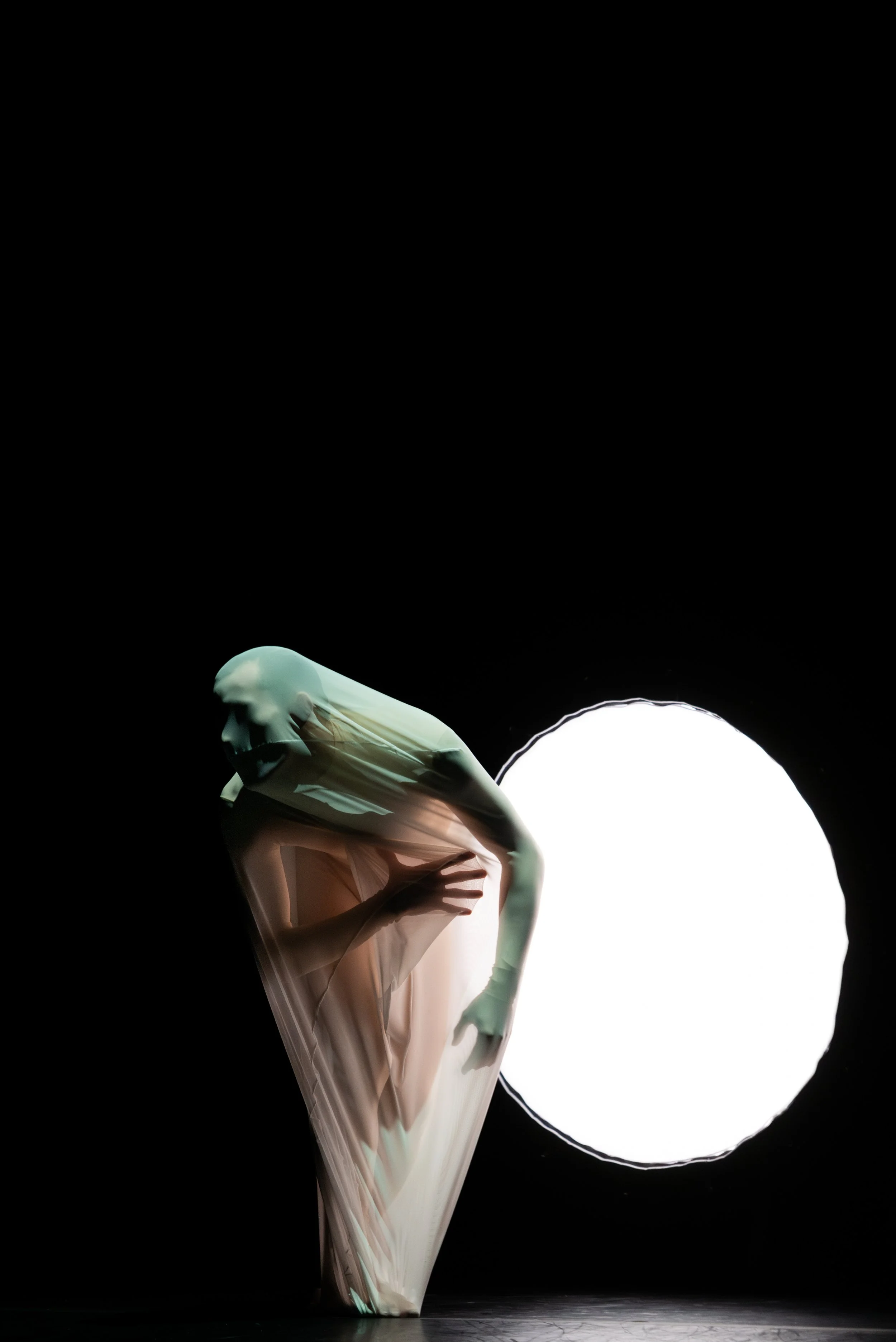 A dancer in a flowing beige dress with long gloves, balancing on one leg with the other bent, near a bright white circular stage light against a dark background.