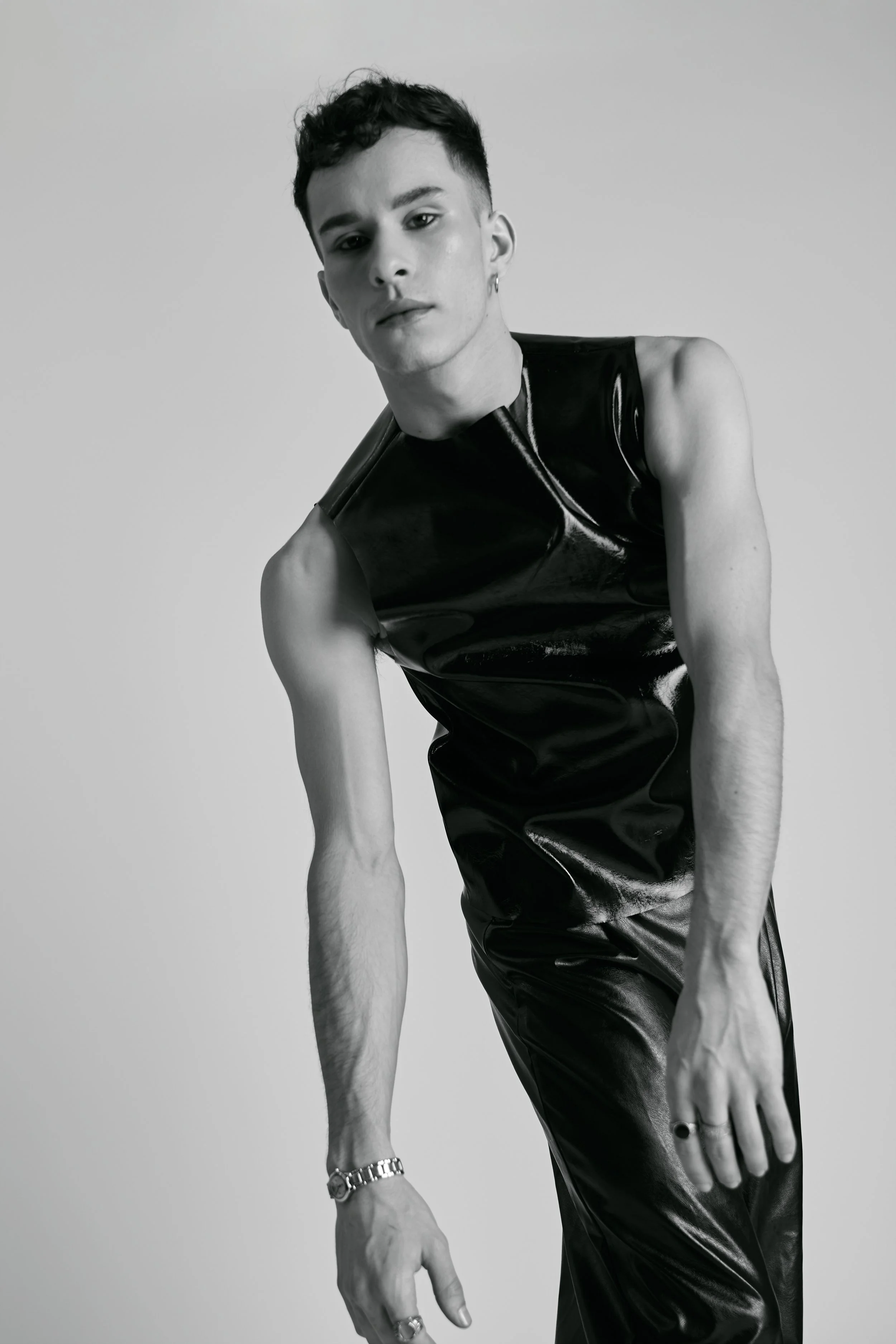 A black-and-white photo of a young person with short dark hair, wearing sleeveless leather-like top, multiple rings, a chain necklace, and a bracelet, posing with a serious expression against a plain background.